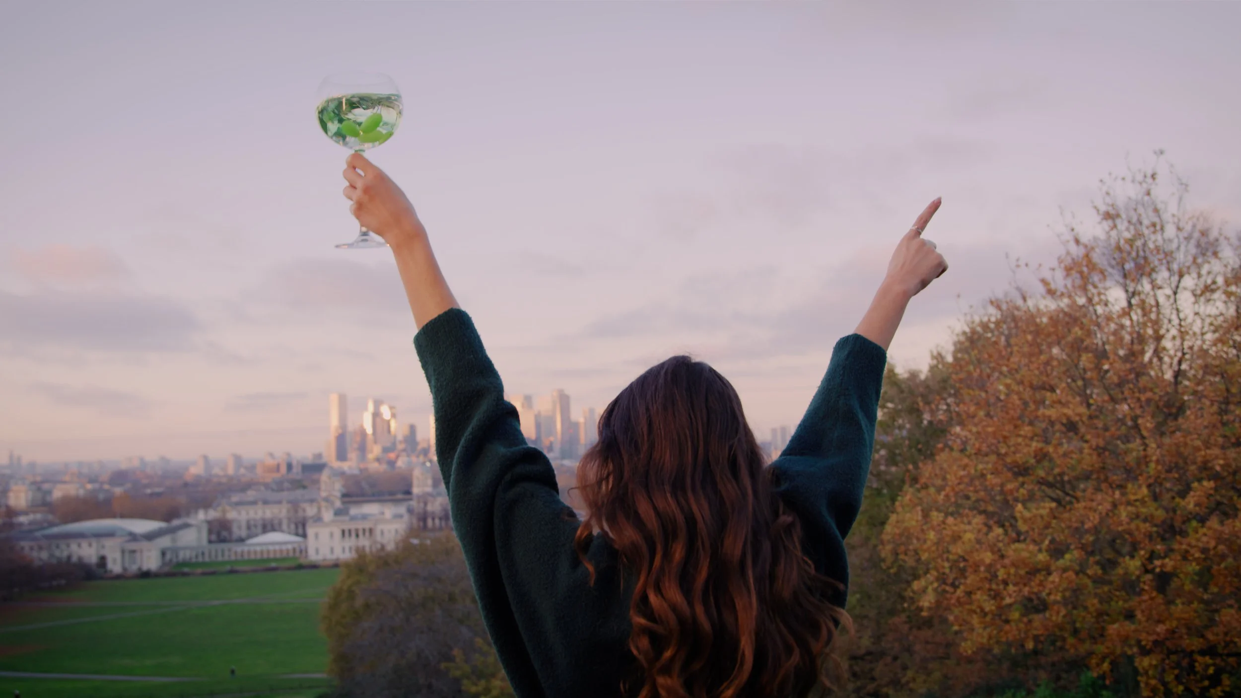 Woman with long, wavy hair raising her arms, holding a glass of wine outdoors with a city skyline and autumn trees in the background.