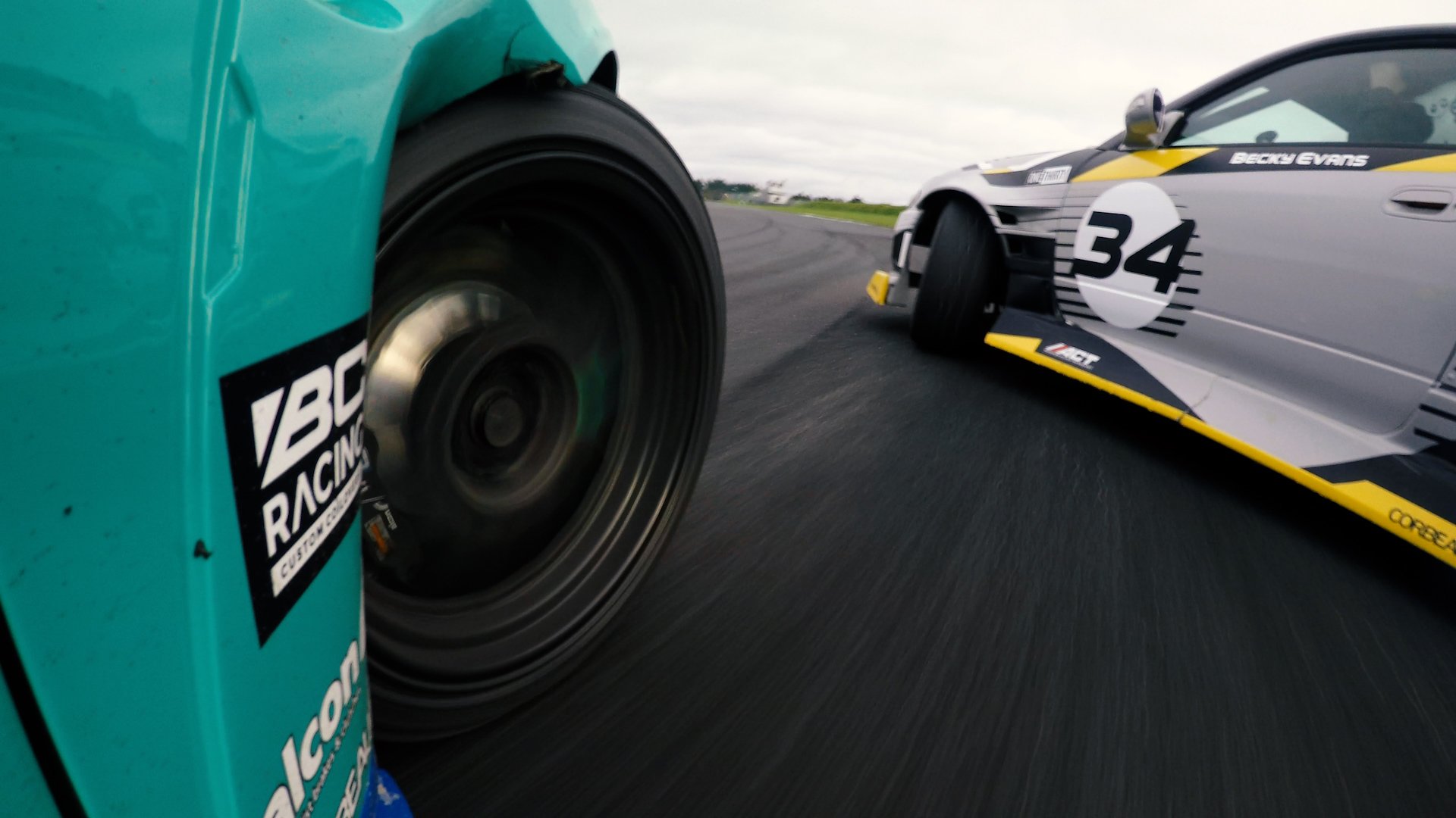 Close-up of two race cars on a track, with the front wheels of a teal car and the side of a silver and yellow car visible, with the number 34 and the name Becky Evans.
