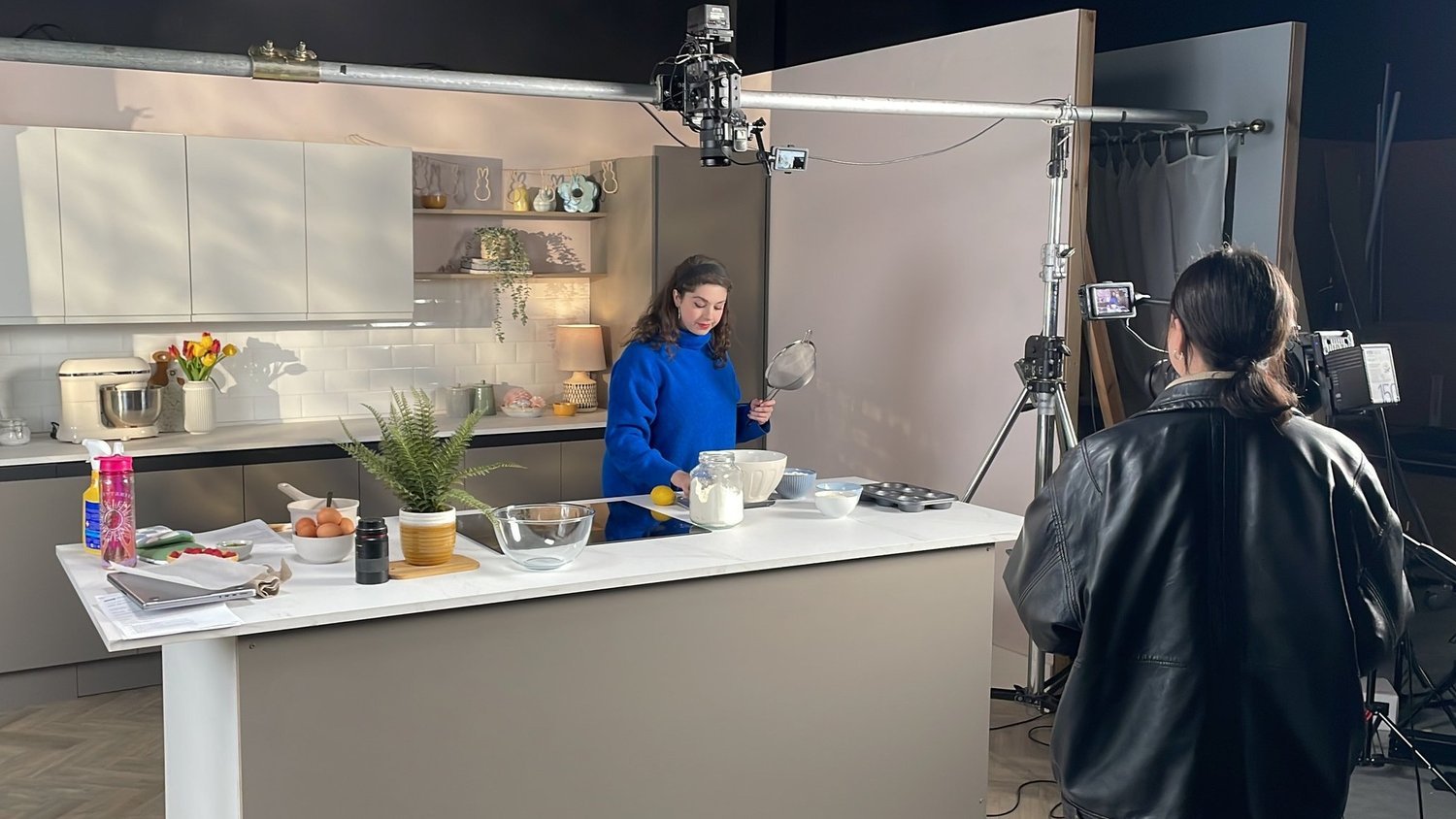 A woman in a blue sweater standing in a kitchen studio set, preparing baking ingredients while a camera operator films her. The kitchen has a white island with eggs, a plant, and baking tools.