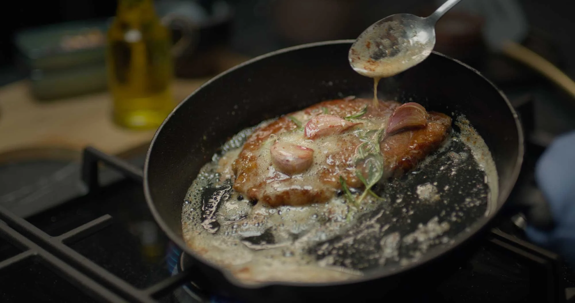 Cooking steak with garlic and herbs in a frying pan on a stove, with liquid sauce being spooned over the meat
