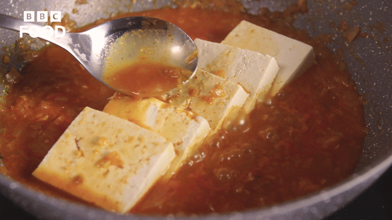 Tofu being cooked in spicy sauce in a frying pan with a spoon.