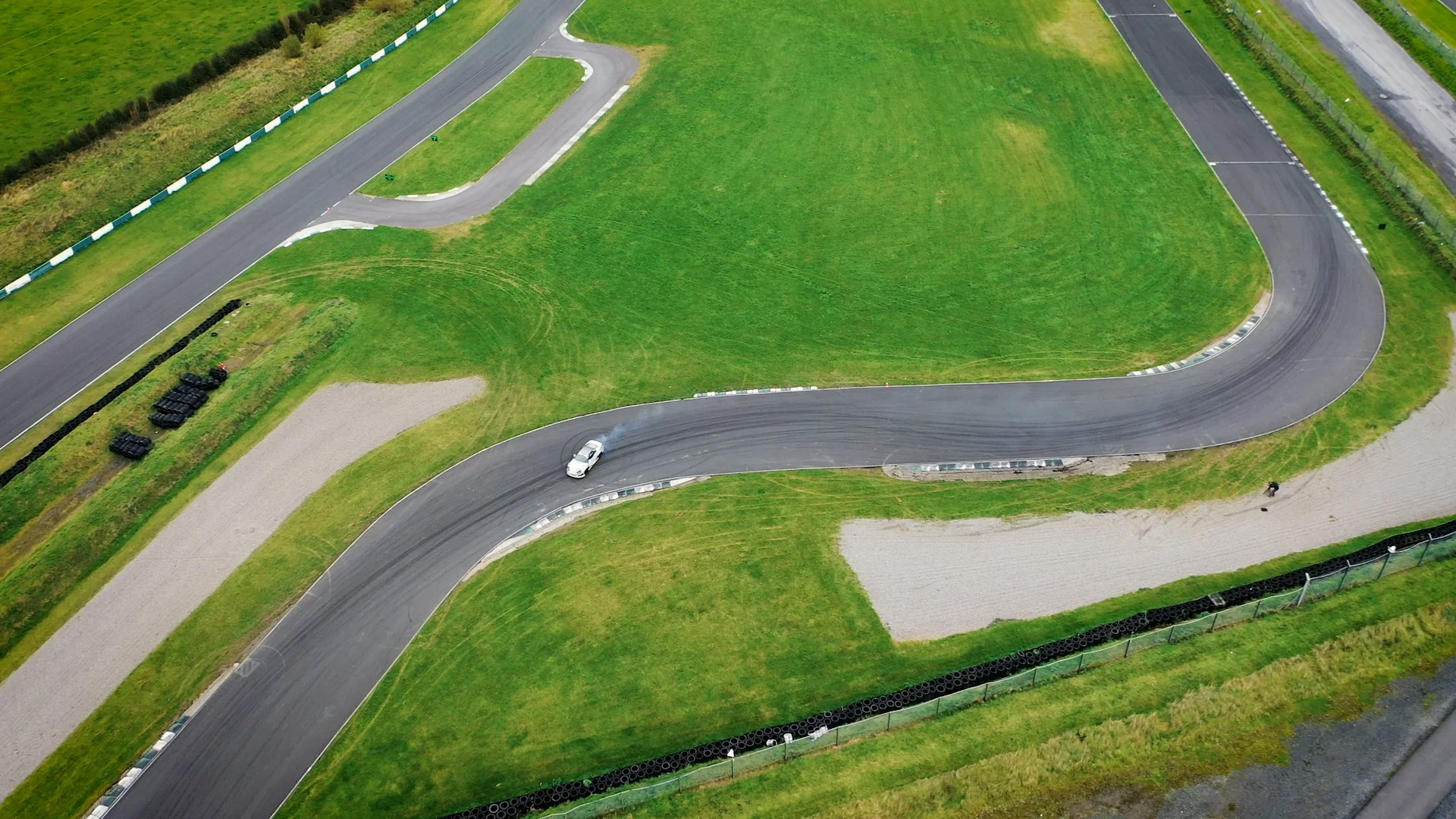 Aerial view of a race track with a single white car on a bend, surrounded by green grass and tire barriers.