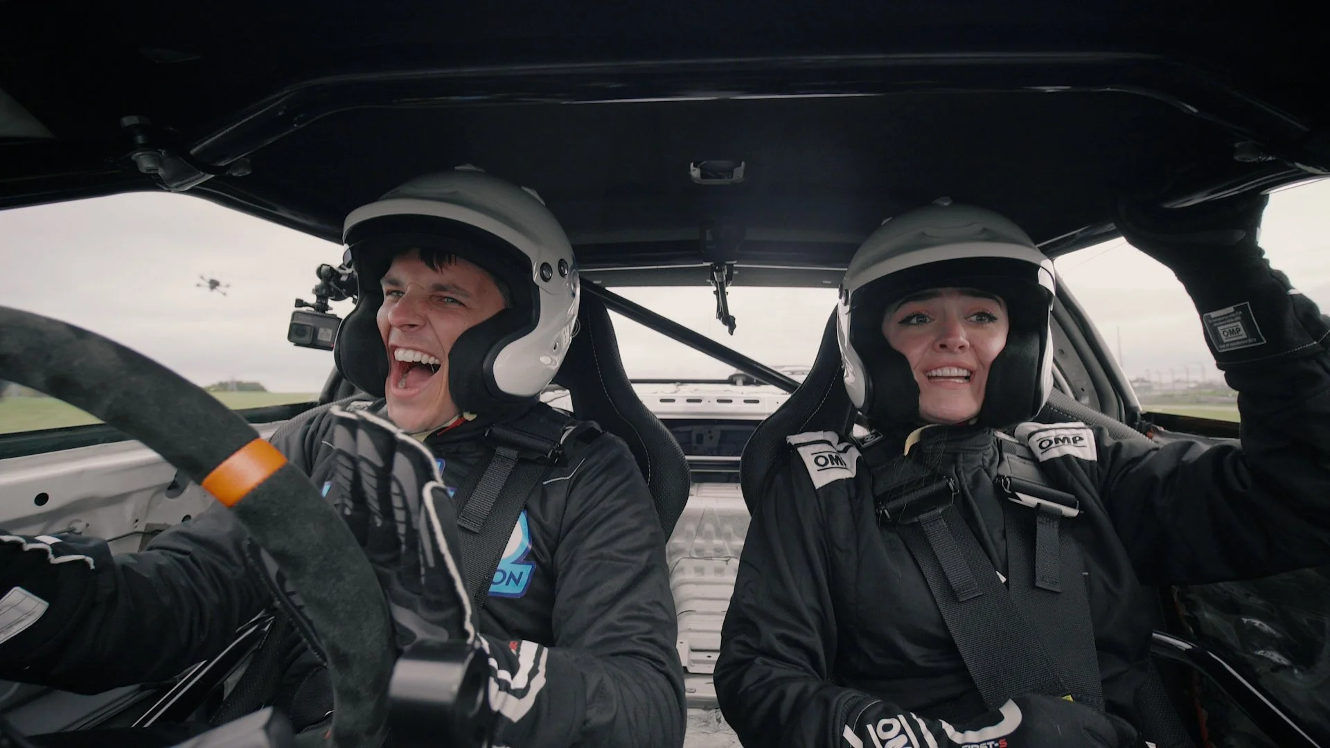 Two race car drivers in racing suits and helmets inside a car, with one smiling widely and the other looking distressed, as a drone flies outside the car on a race track.