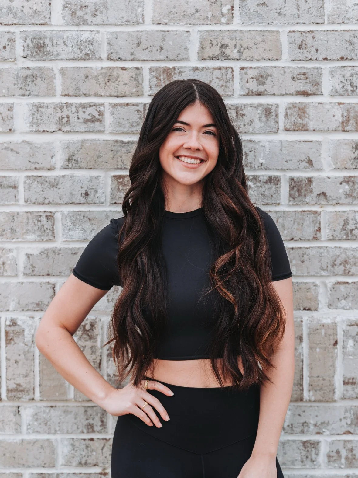 A young woman with long dark hair smiling and standing outdoors on a brick patio, surrounded by green bushes and trees. She's wearing a white lace blouse, denim shorts, and pink sneakers. She has one hand behind her head and is holding a small striped bag in her other hand.