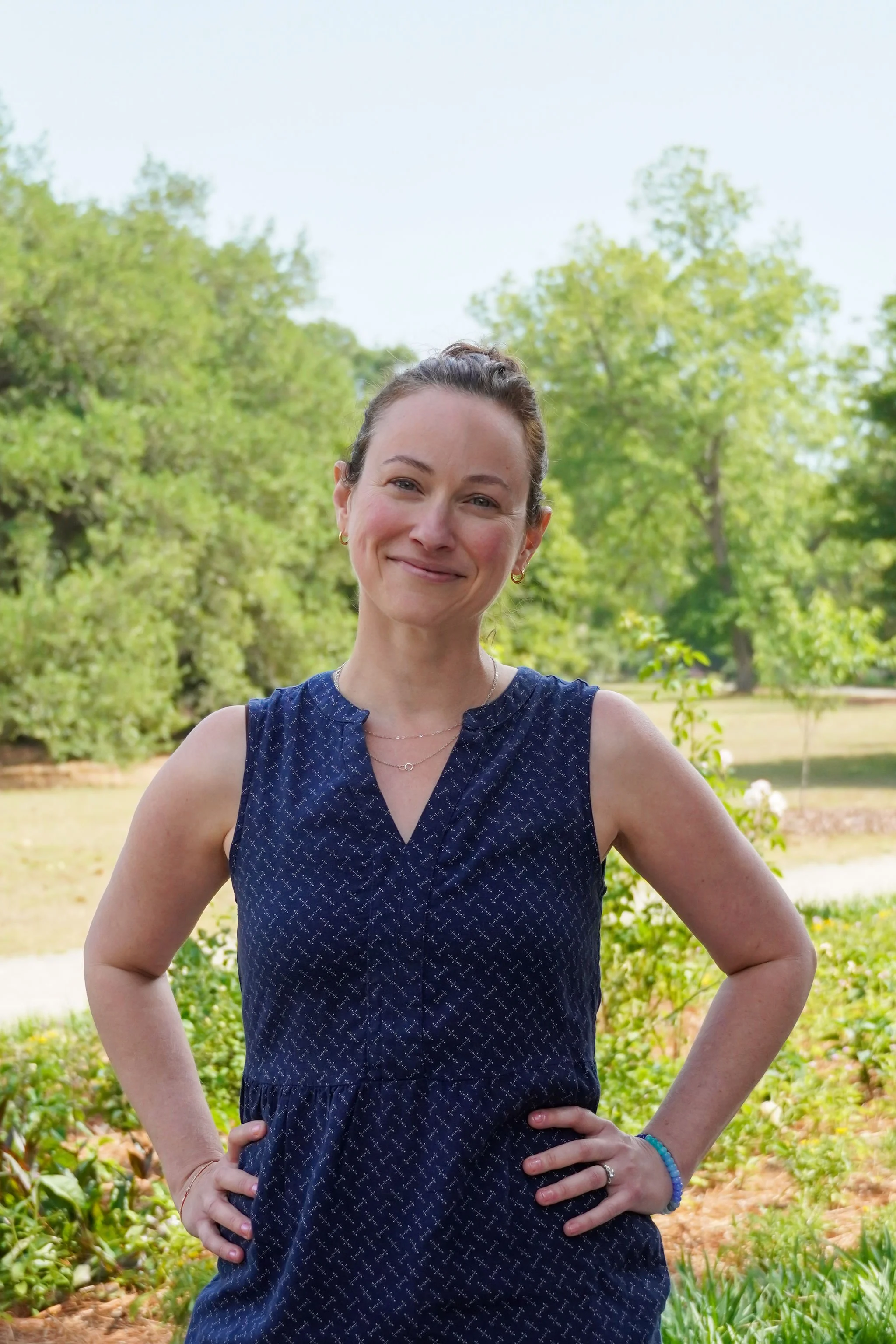 Young woman in a blue dress posing in a park and smiling with hands on her hips