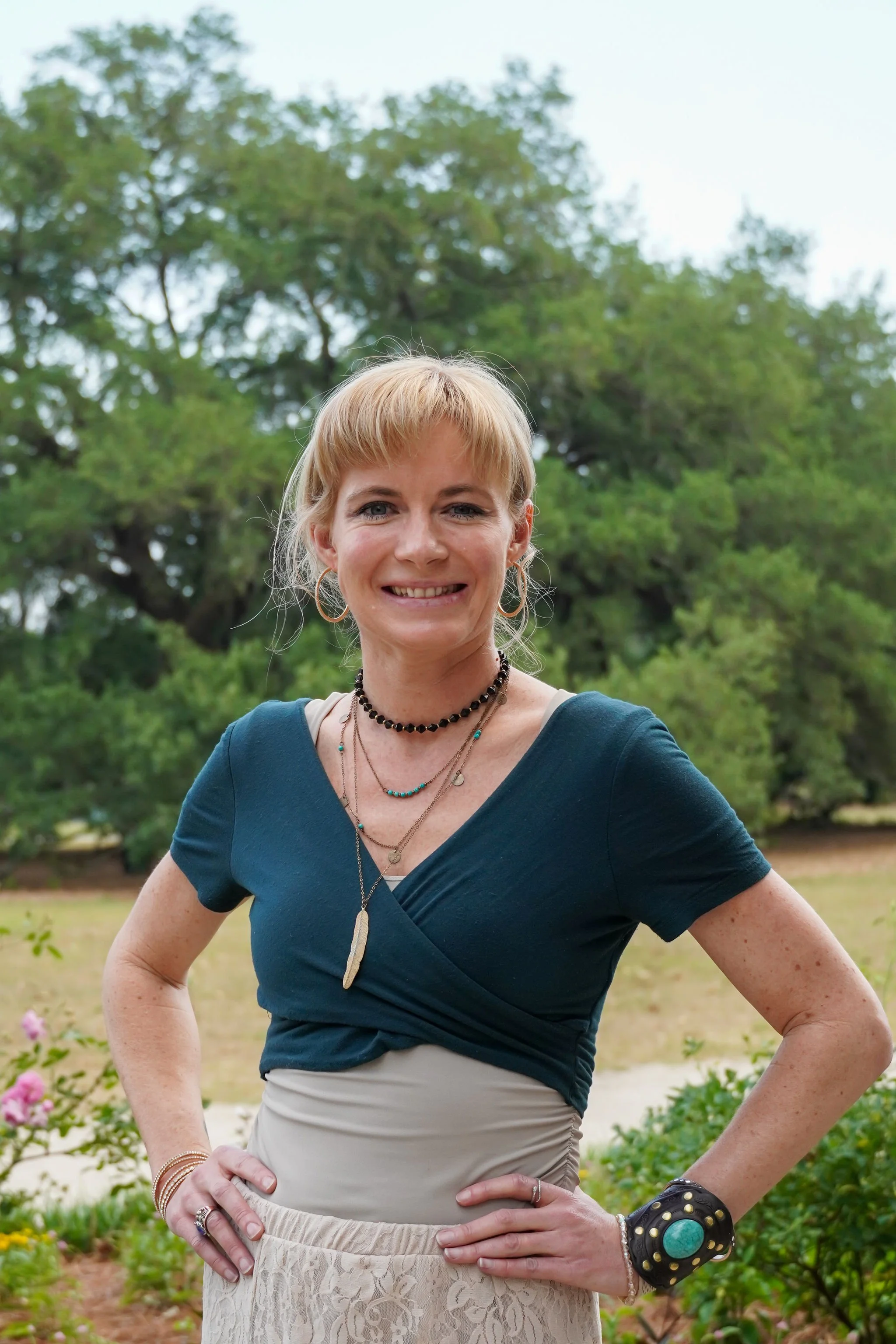 Young woman in teal top posing with her hands on her hips and smiling at a park