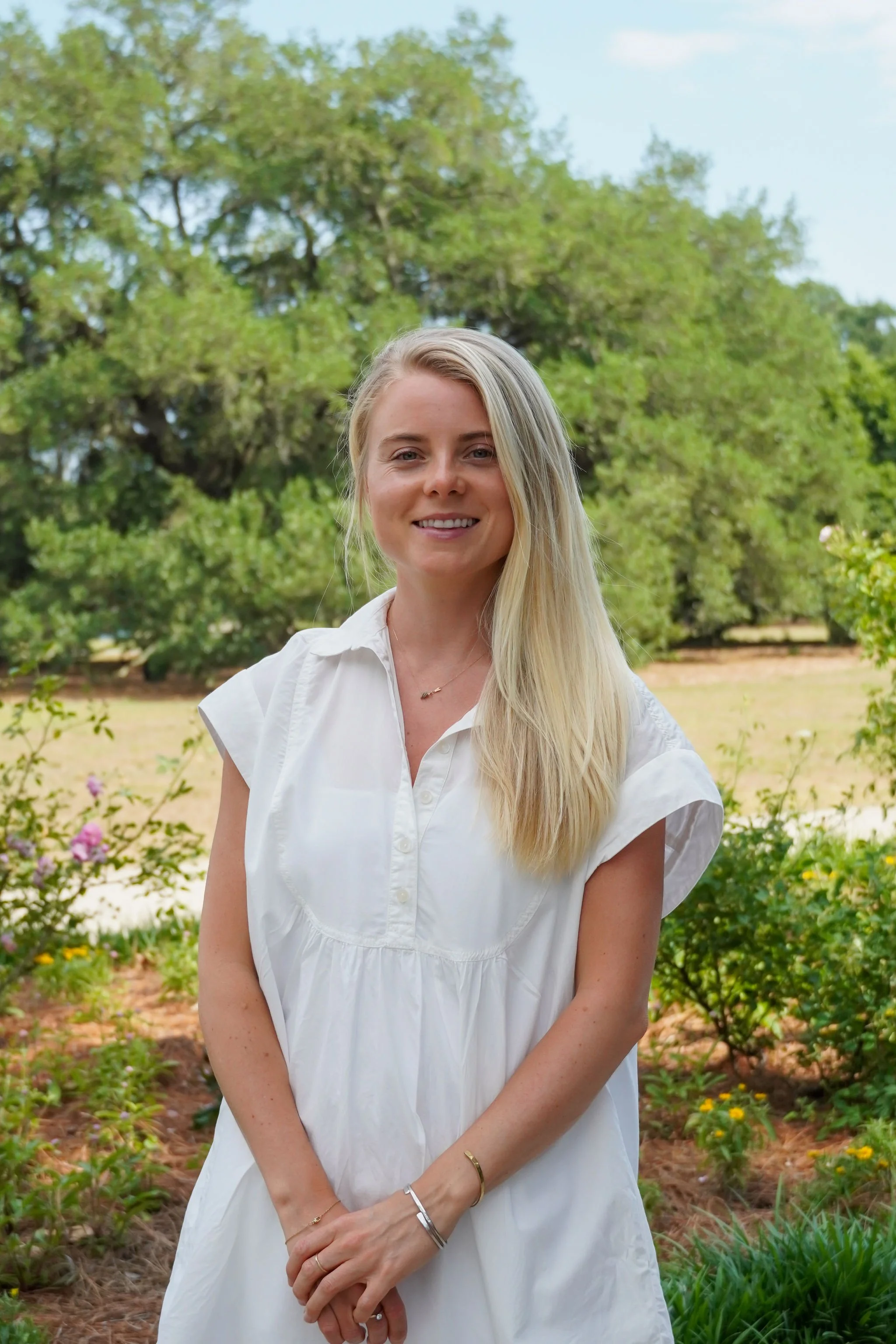 Young woman in a white dress posing in a park, smiling and holding her hands