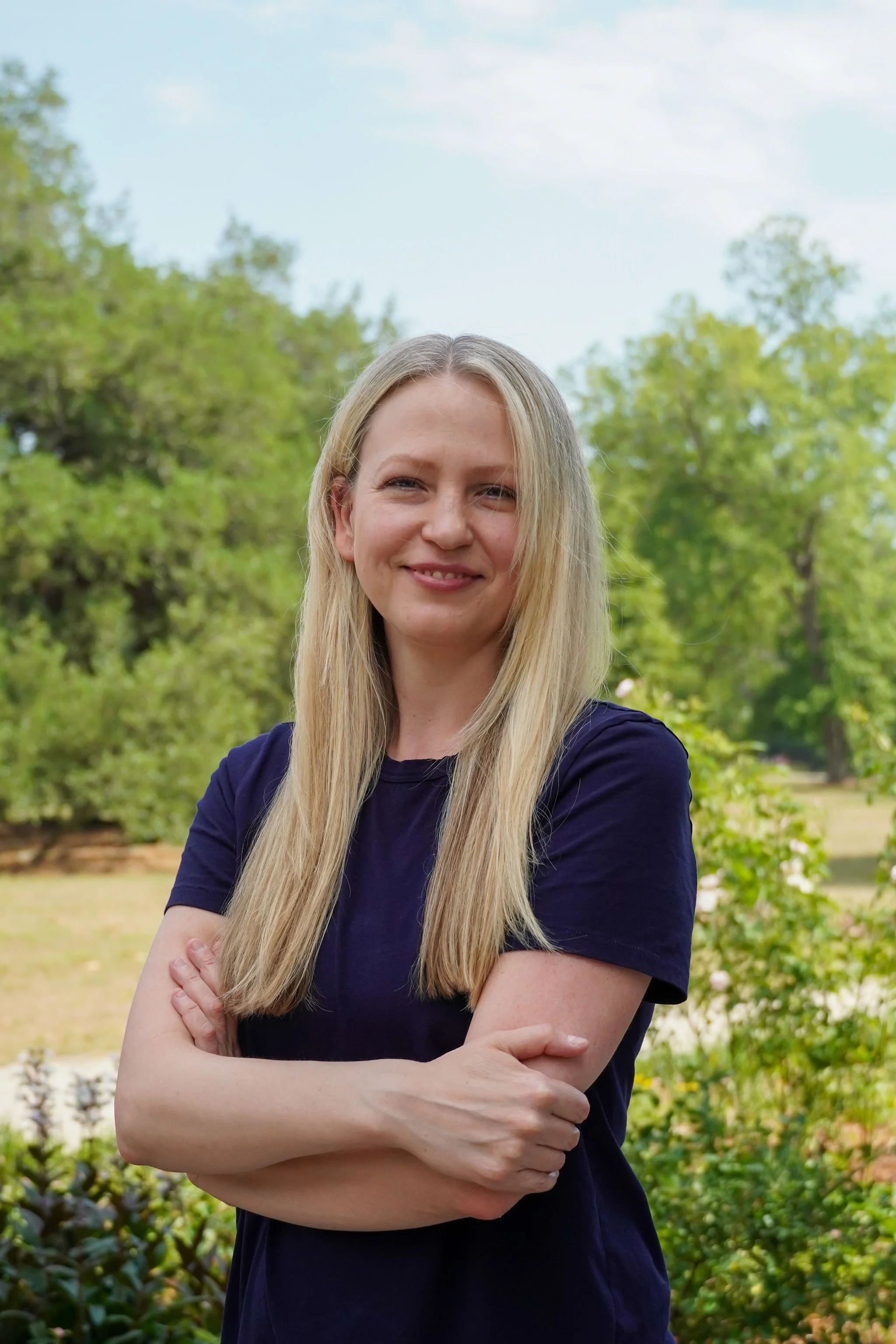 Young woman in a navy blue shirt posing in a park with arms crossed and smiling