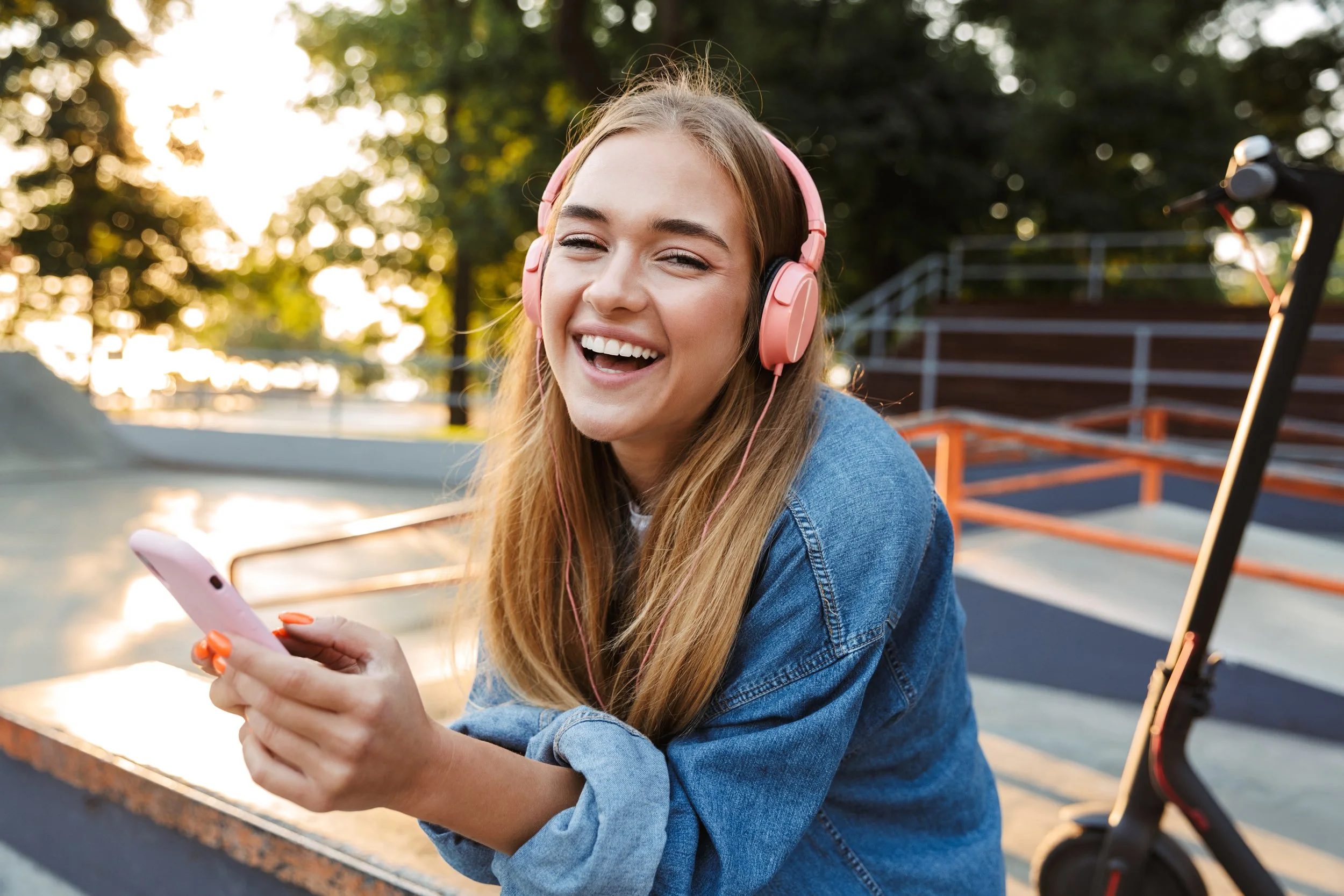 Young woman with long blonde hair wearing pink headphones, smiling while looking at her smartphone, sitting at a skate park outdoors during sunset with skate ramps and trees in the background.