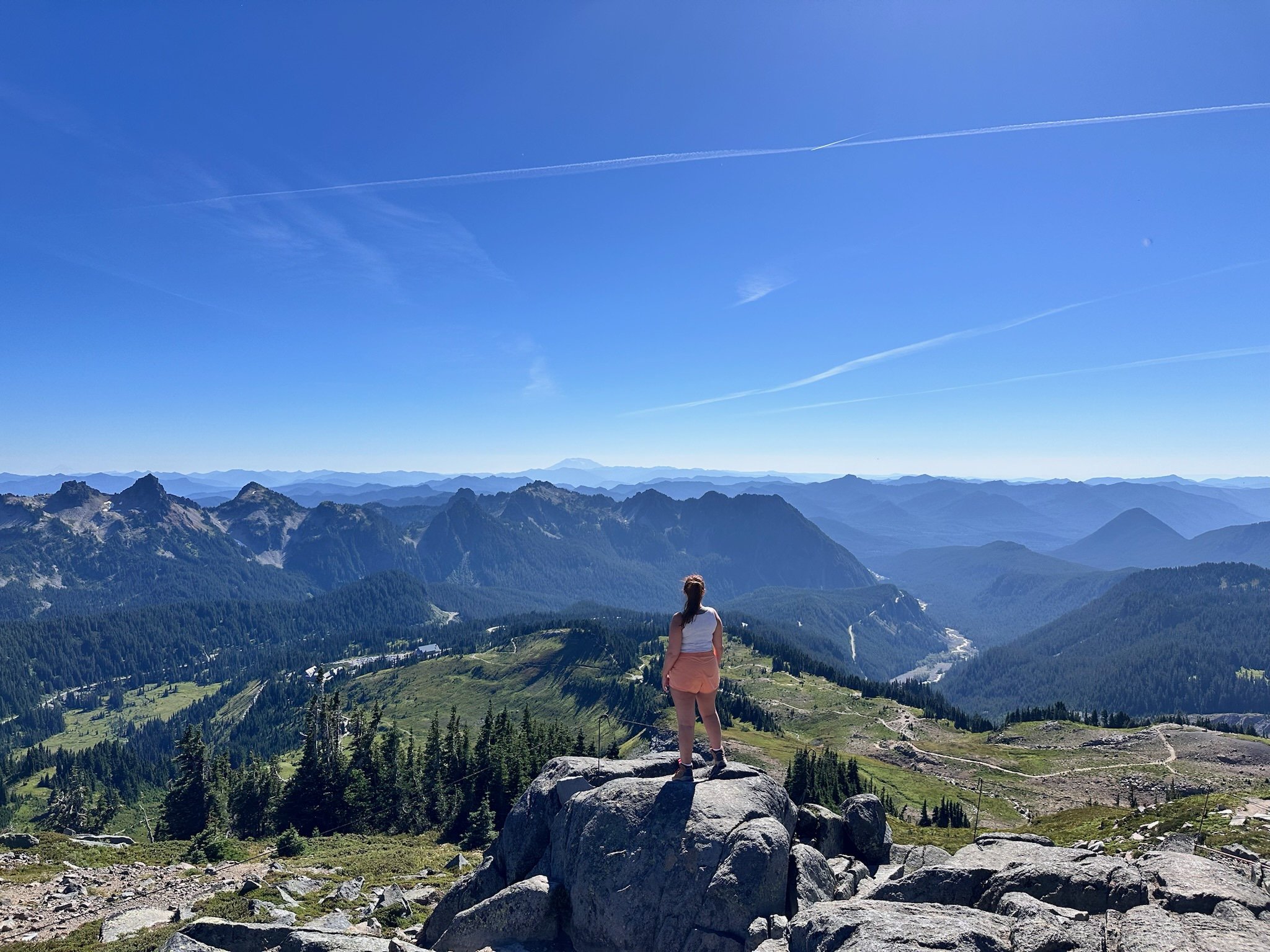 A woman standing on a large rock, overlooking a vast mountain range under a clear blue sky.