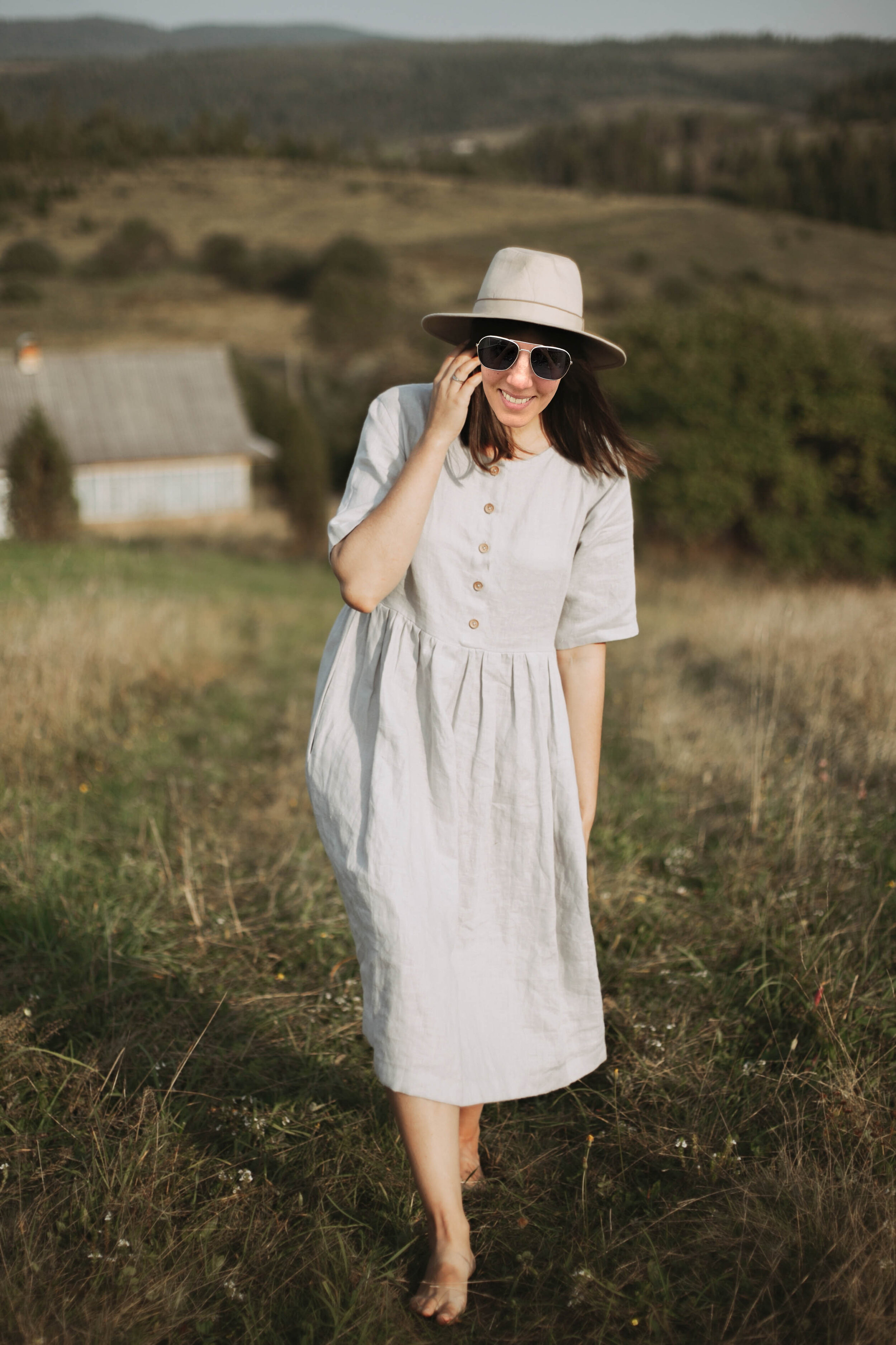 Stylish girl in linen dress and hat walking barefoot in grass in sunny field at village. Boho woman relaxing in countryside, 