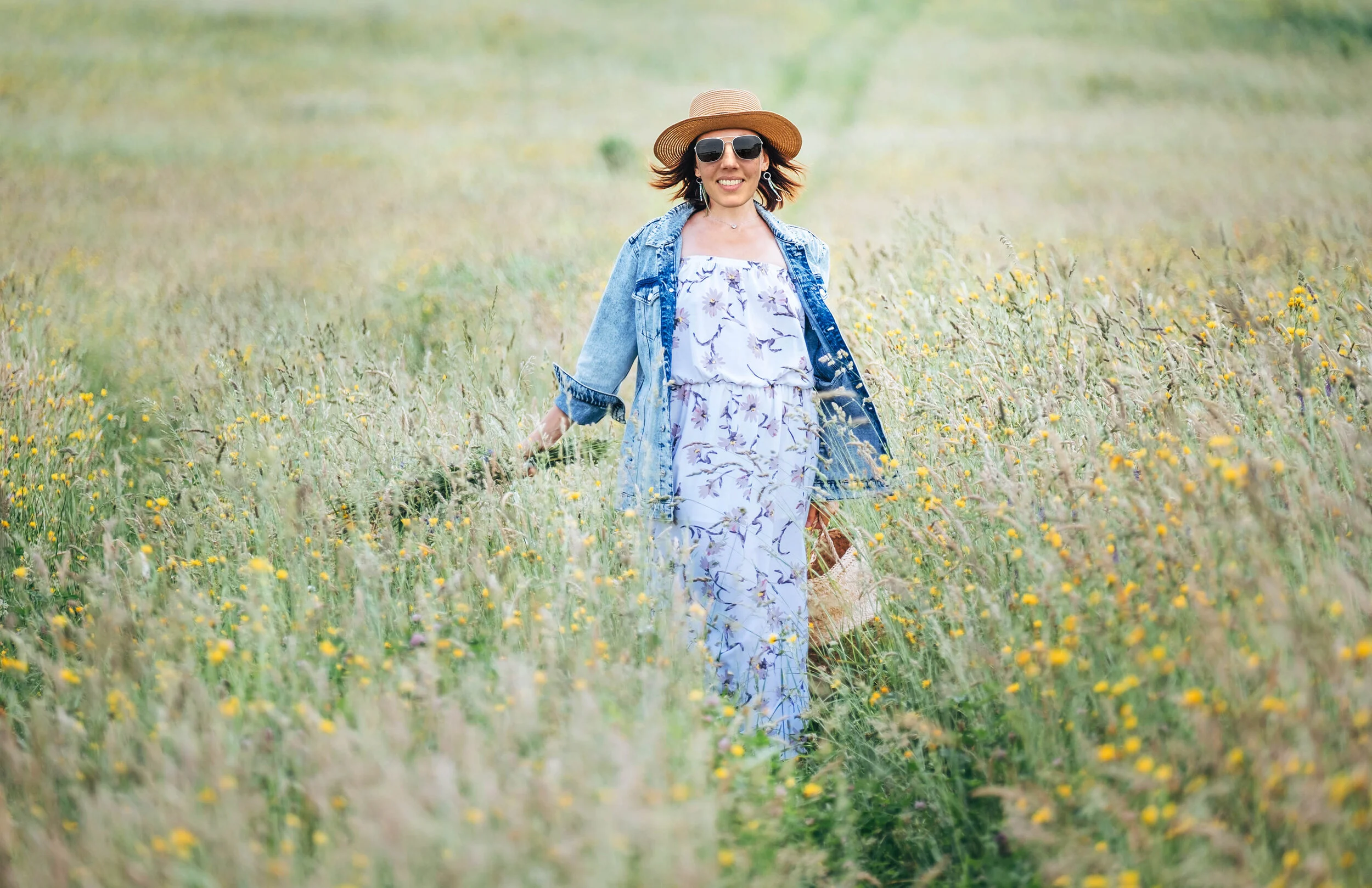 Sincerely smiling young woman dressed jeans jacket and light summer dress walking by the high green grass meadow with basket and