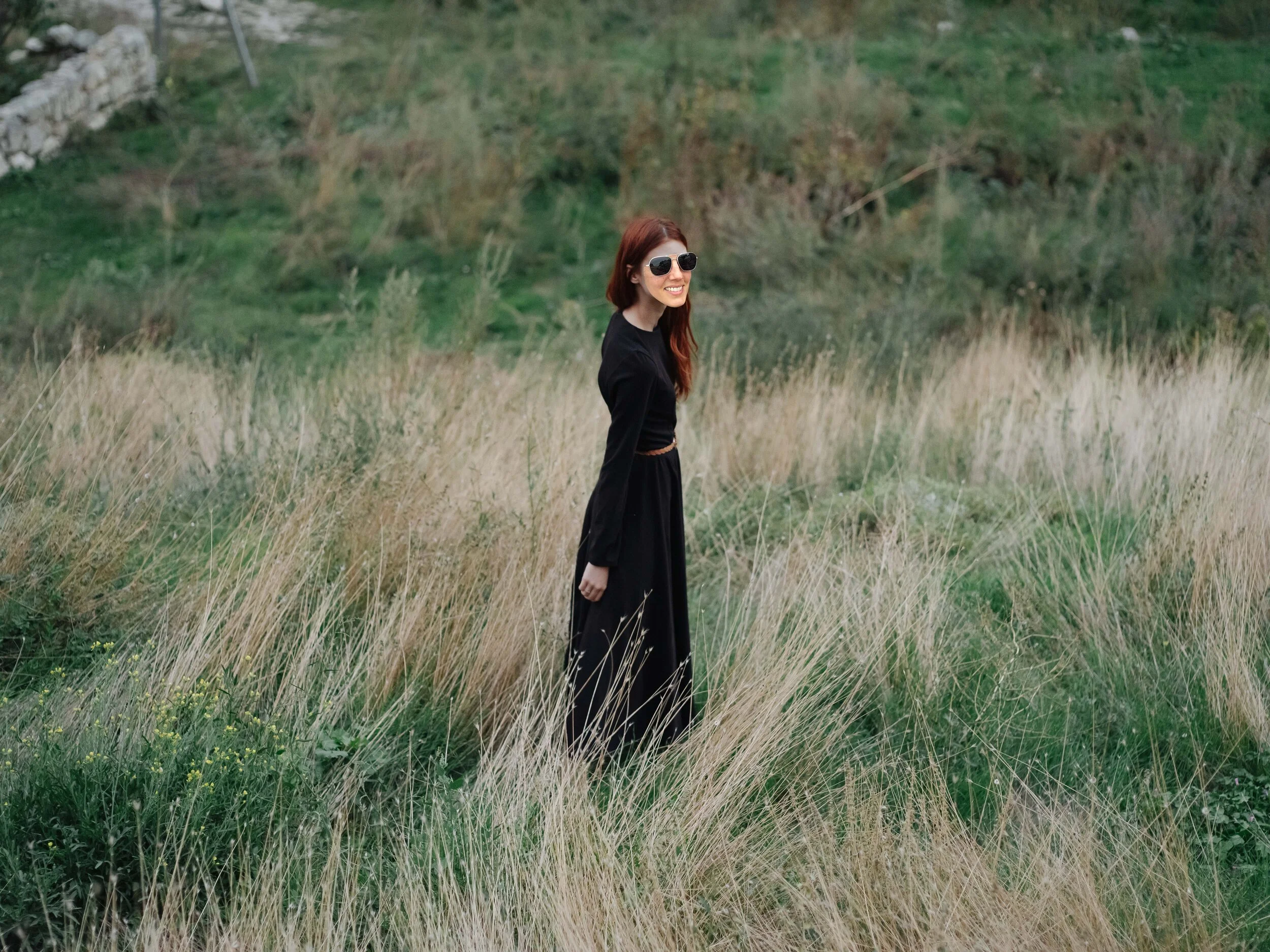 You are a young woman in a black dress walking on dry grass in a meadow in the mountains to nature