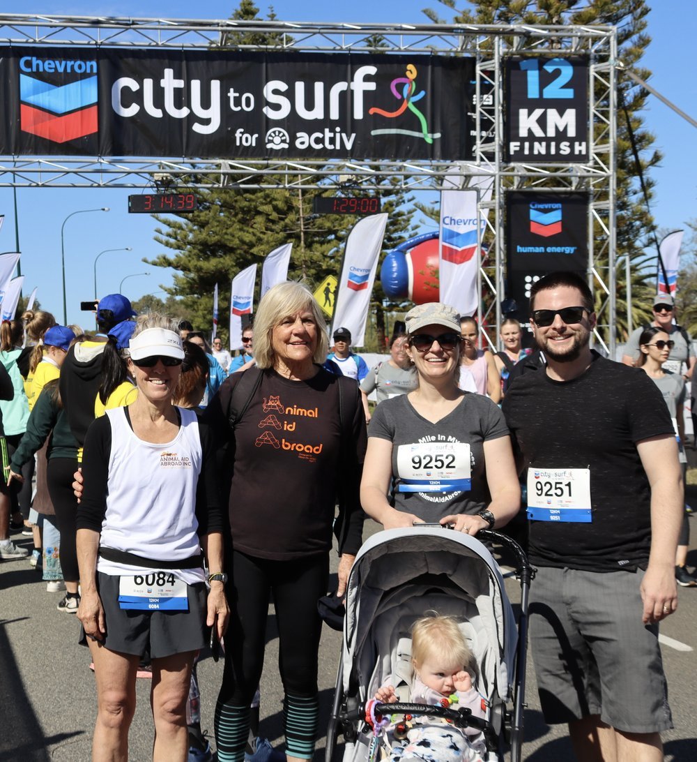 Animal Aid Abroad Members and Volunteers at the finish line of a 12 km race, with a banner reading 'city to surf for activ' overhead, and other race participants in the background.