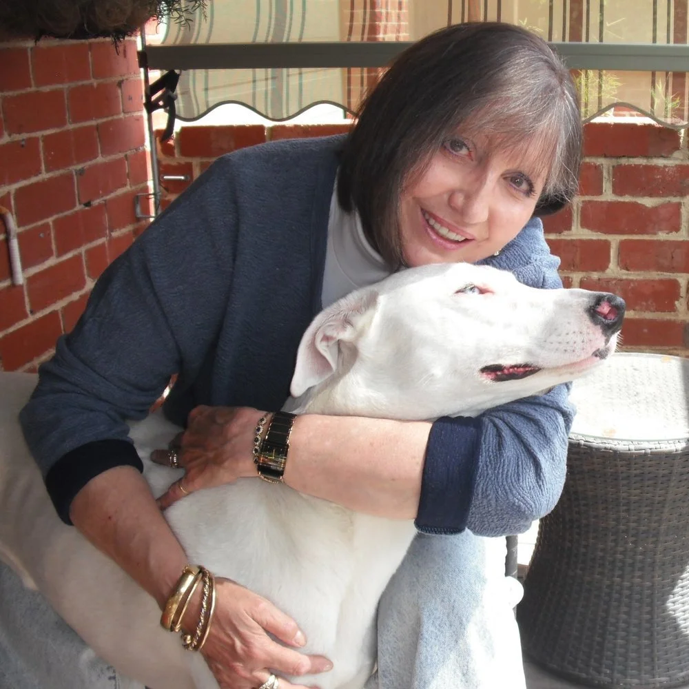 A woman with shoulder-length gray hair smiling and hugging a white dog hanging out, sitting on a patio with brick walls and a wicker chair in the background.