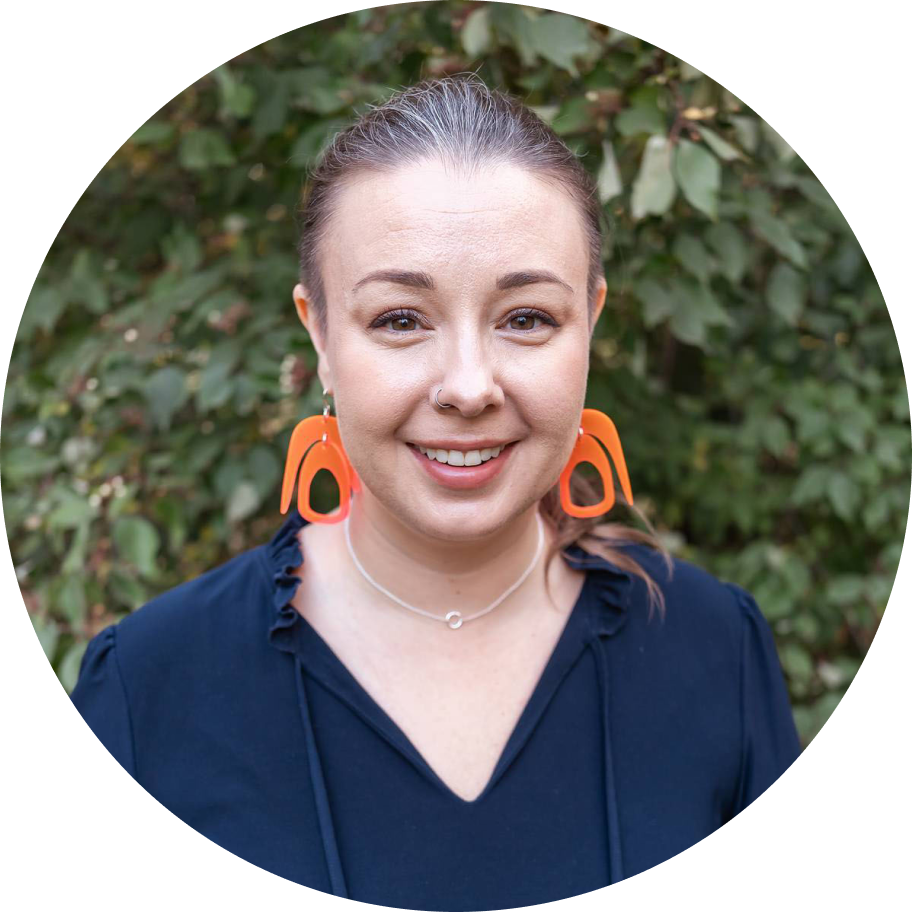 A close-up portrait of a smiling white woman with brown hair, a nose ring, and vibrant orange geometric earrings, framed by green leaves.