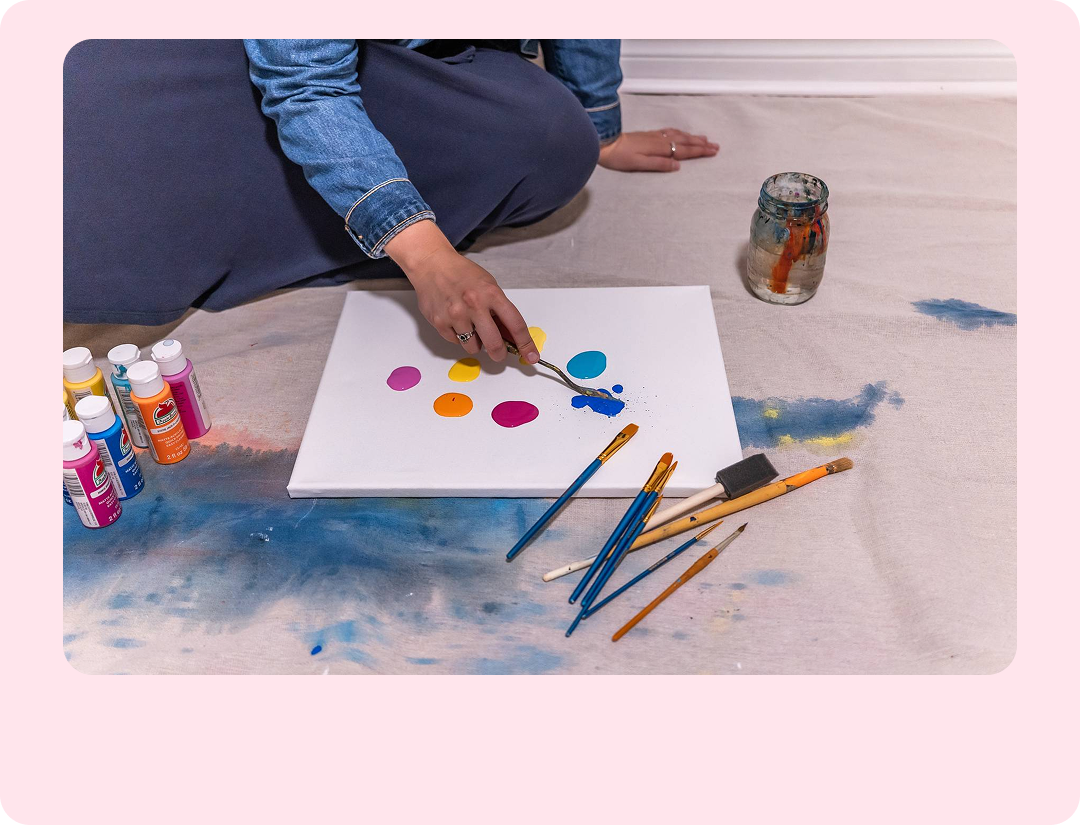 A person in a denim jacket and dark skirt sits cross-legged on the floor, using a palette knife to mix colorful acrylic paint droplets on a white canvas.