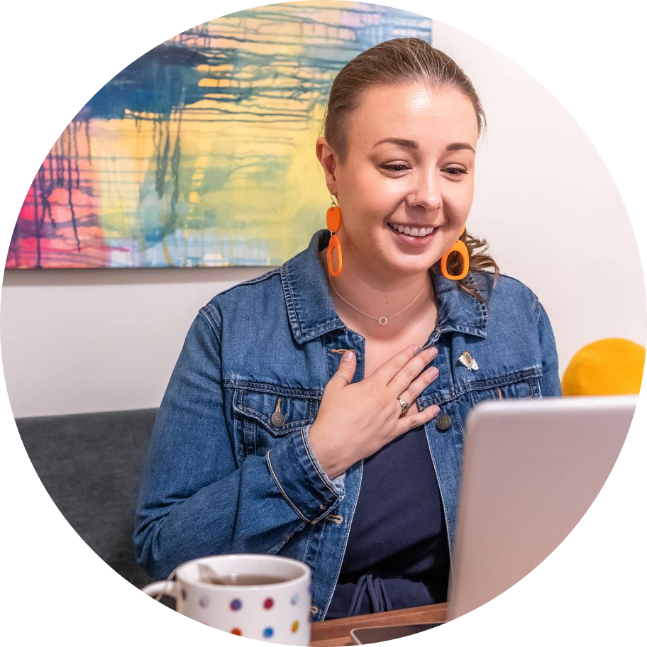 A light-skinned woman with tied-back brown hair, wearing a denim jacket and bright orange earrings, smiles warmly at a laptop with her hand over her heart.