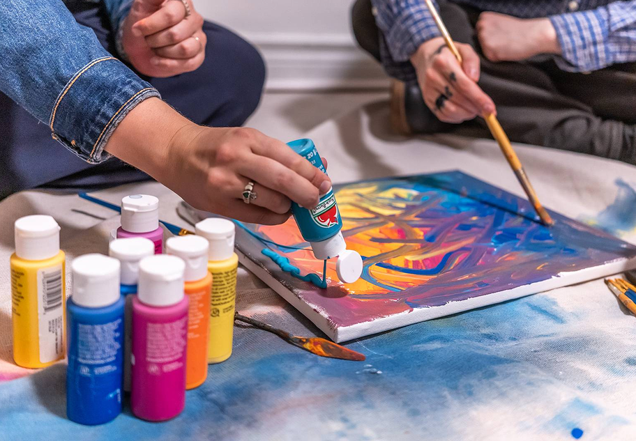 A light-skinned person in a denim jacket squeezes teal paint onto an abstract canvas, sitting beside another person in a plaid shirt painting with a brush.