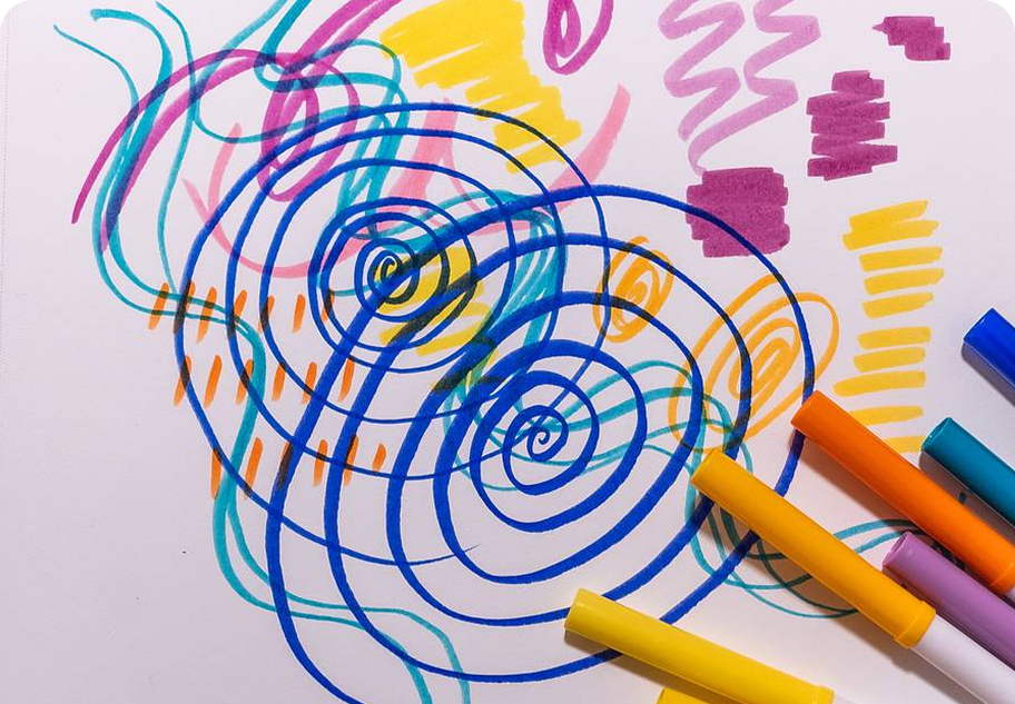Bright blue spiral scribbles and colorful marker patterns on white paper, surrounded by yellow, orange, and purple felt-tip markers.