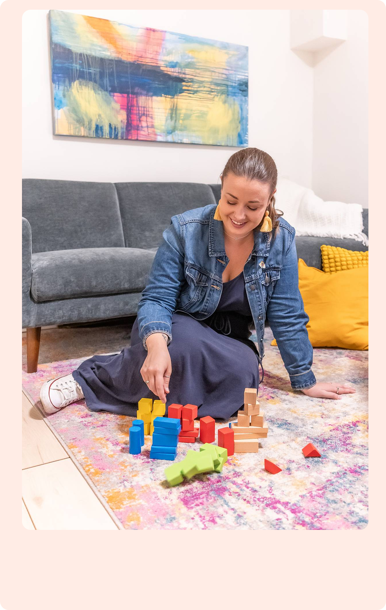 A smiling light-skinned woman with brown hair in a ponytail, wearing a denim jacket and dark dress, sits on a colorful rug arranging wooden blocks.