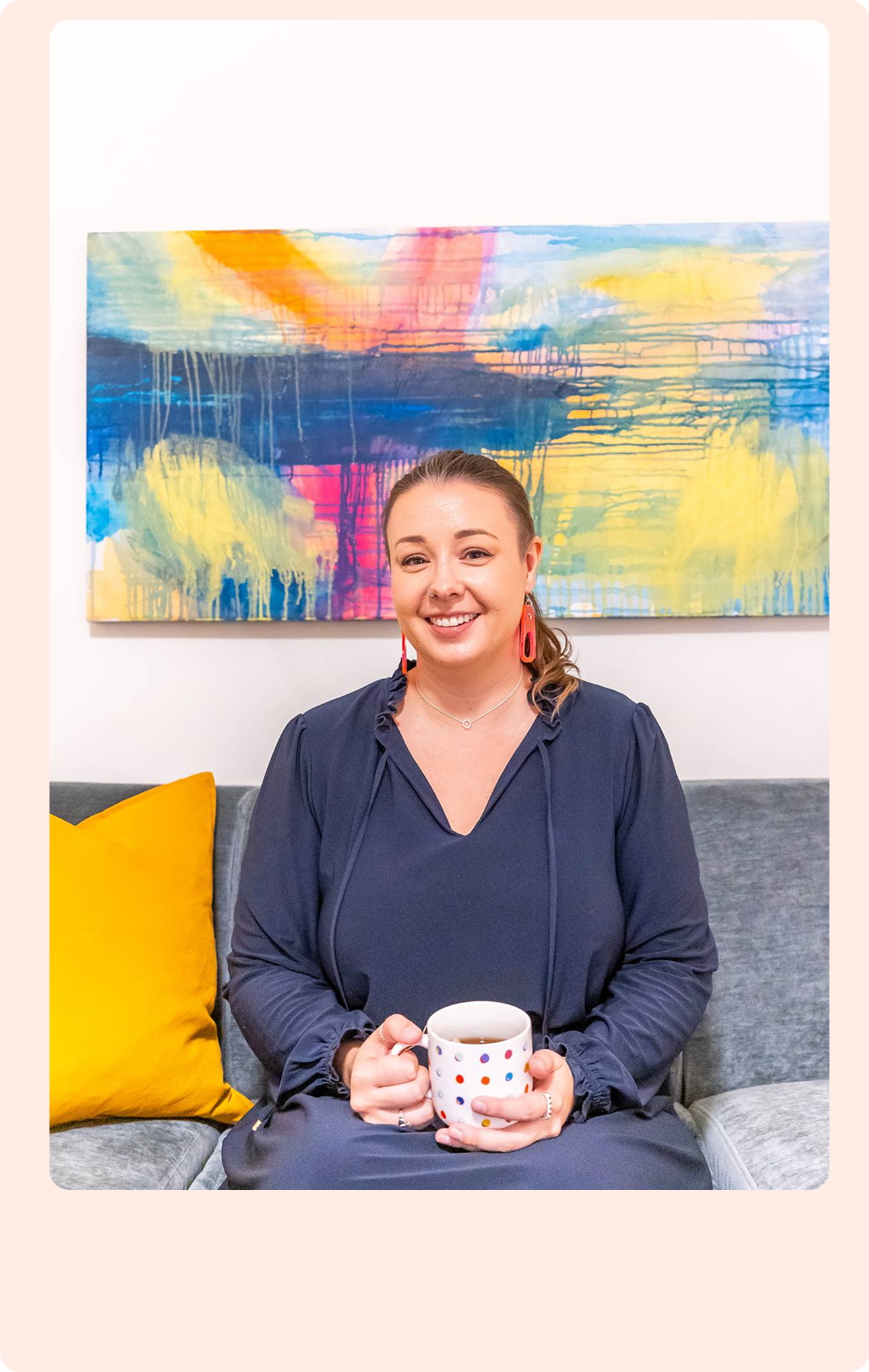 A light-skinned woman with brown hair, wearing a dark dress, orange statement earrings, and a delicate necklace, sits on a sofa smiling with a polka-dot mug and bold colourful wall painting.