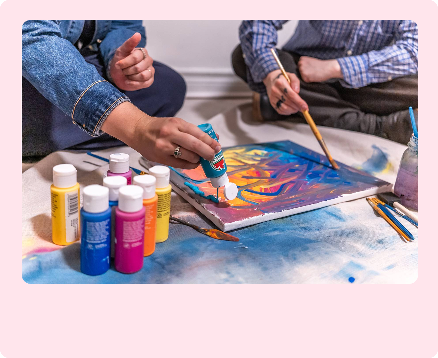 A light-skinned person in a denim jacket squeezes teal paint onto an abstract canvas, sitting beside another person in a plaid shirt painting with a brush.