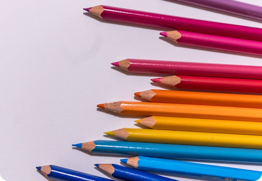 Sharpened colored pencils in rainbow order—pinks, reds, oranges, yellows, and blues—lay arranged horizontally across a white background.