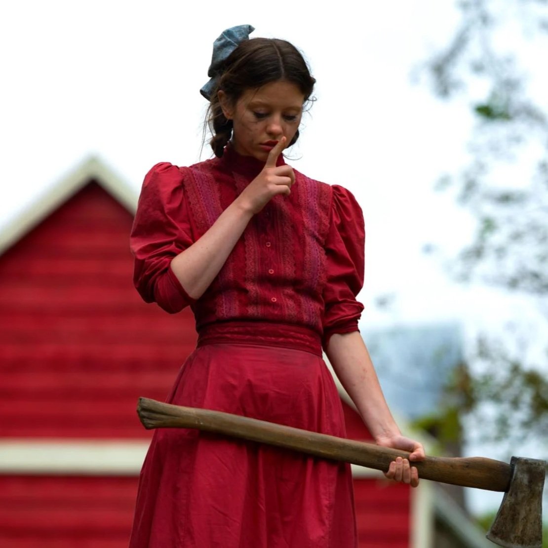 Still from the movie, pearl: Woman holding an axe looking down with a finger on her mouth shushing 