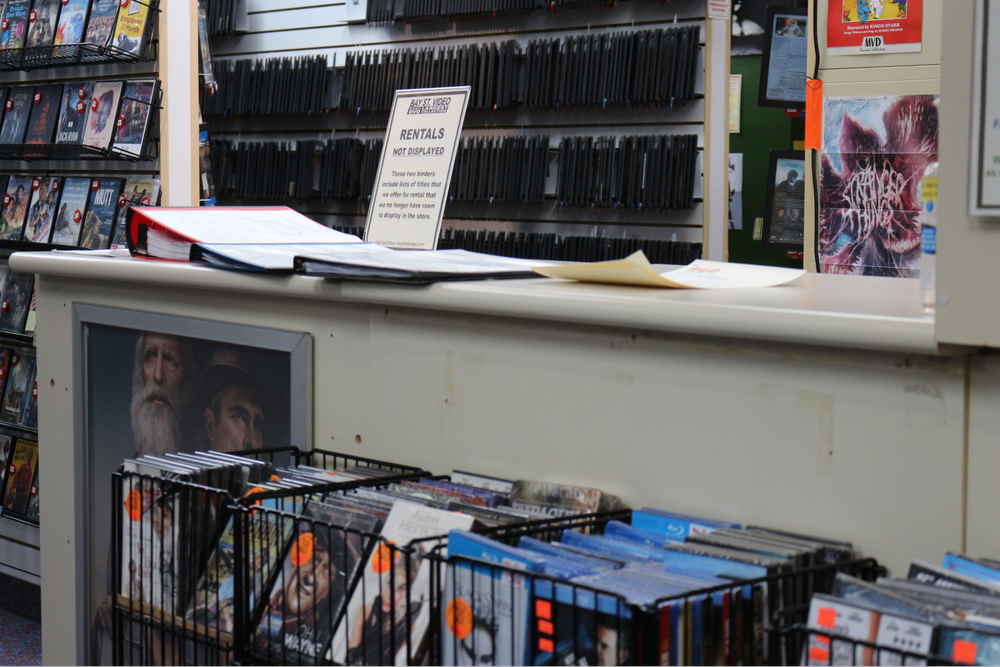  Racks of movies are set below a customer counter with binders on it. 