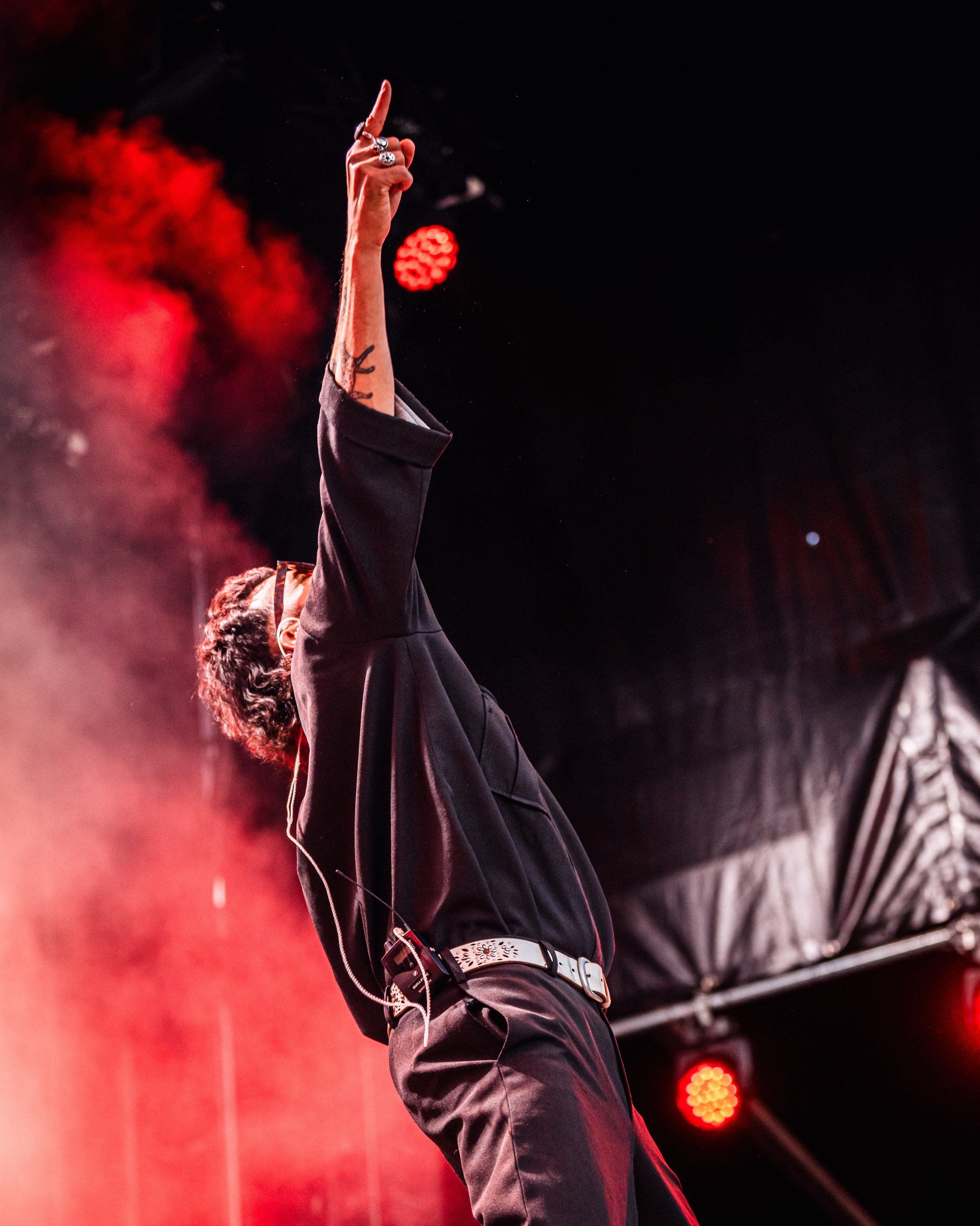 Un musicien sur scène, levant le bras avec le doigt pointé en l'air, dans une ambiance de concert avec des lumières rouges et de la fumée.