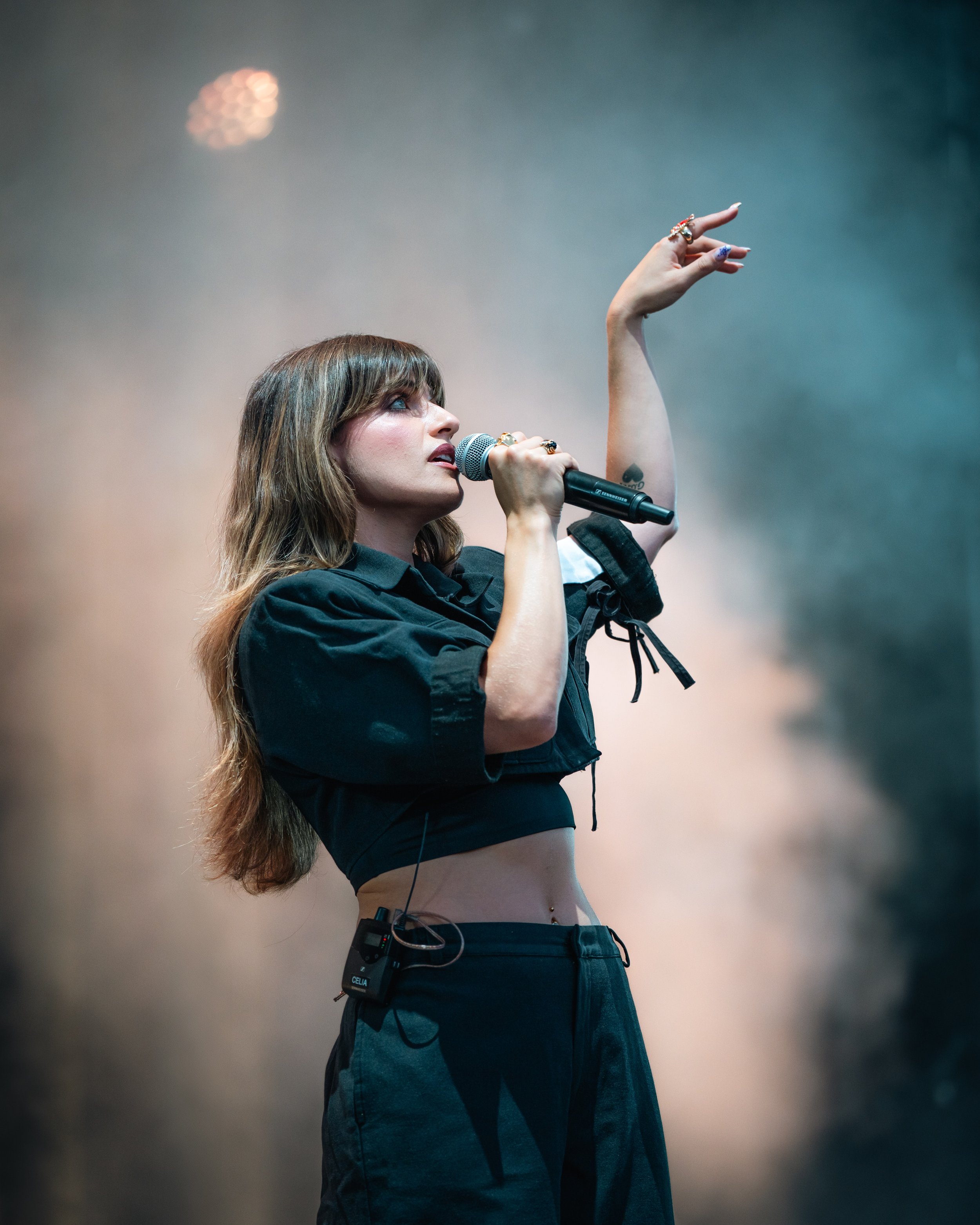 Jeune femme chanteuse avec un microphone, portant un haut court noir, avec un earpiece audio, sur scène avec une lumière colorée en arrière-plan.