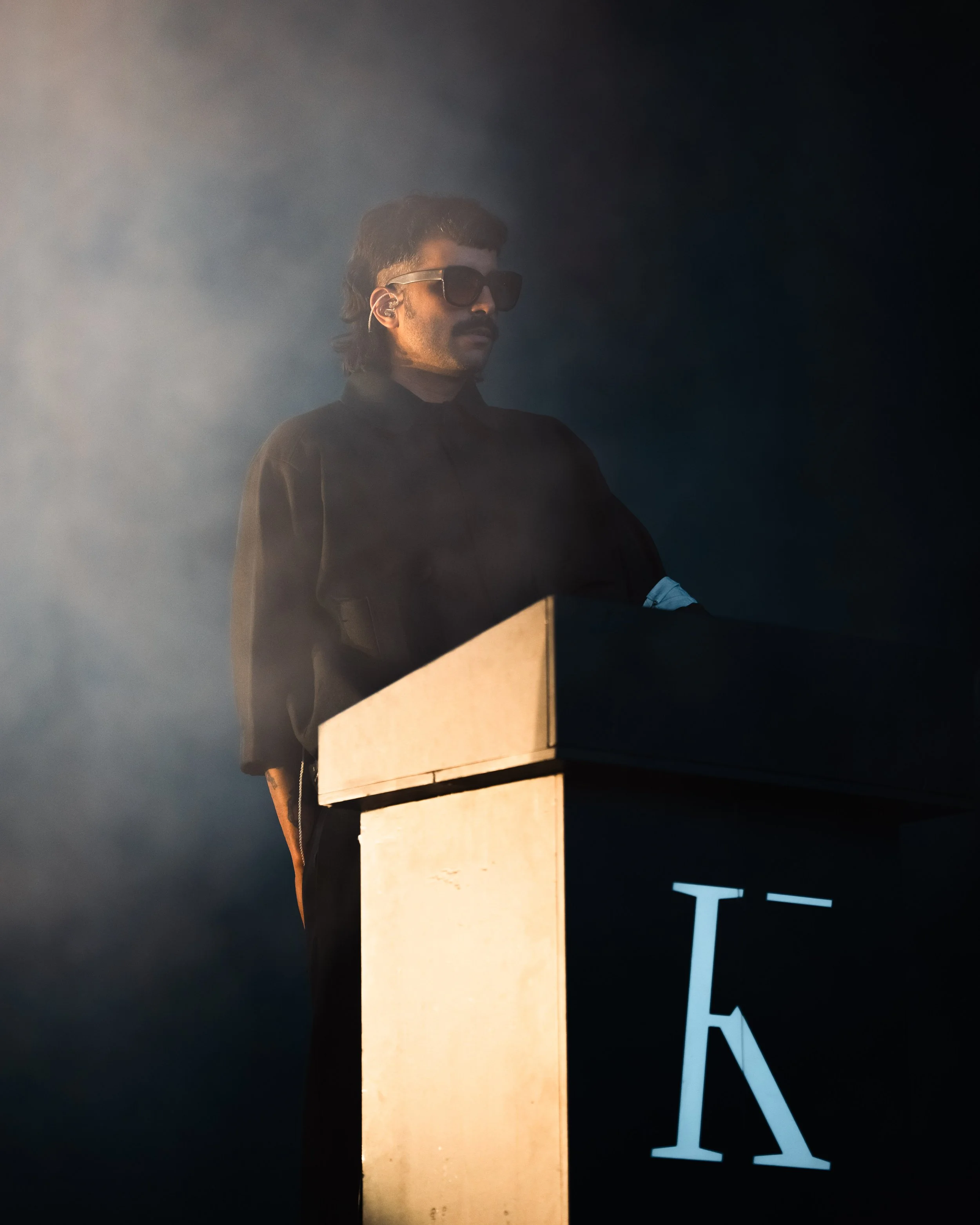 Un homme portant des lunettes de soleil et une veste noire joue du piano dans un environnement sombre avec de la brume.