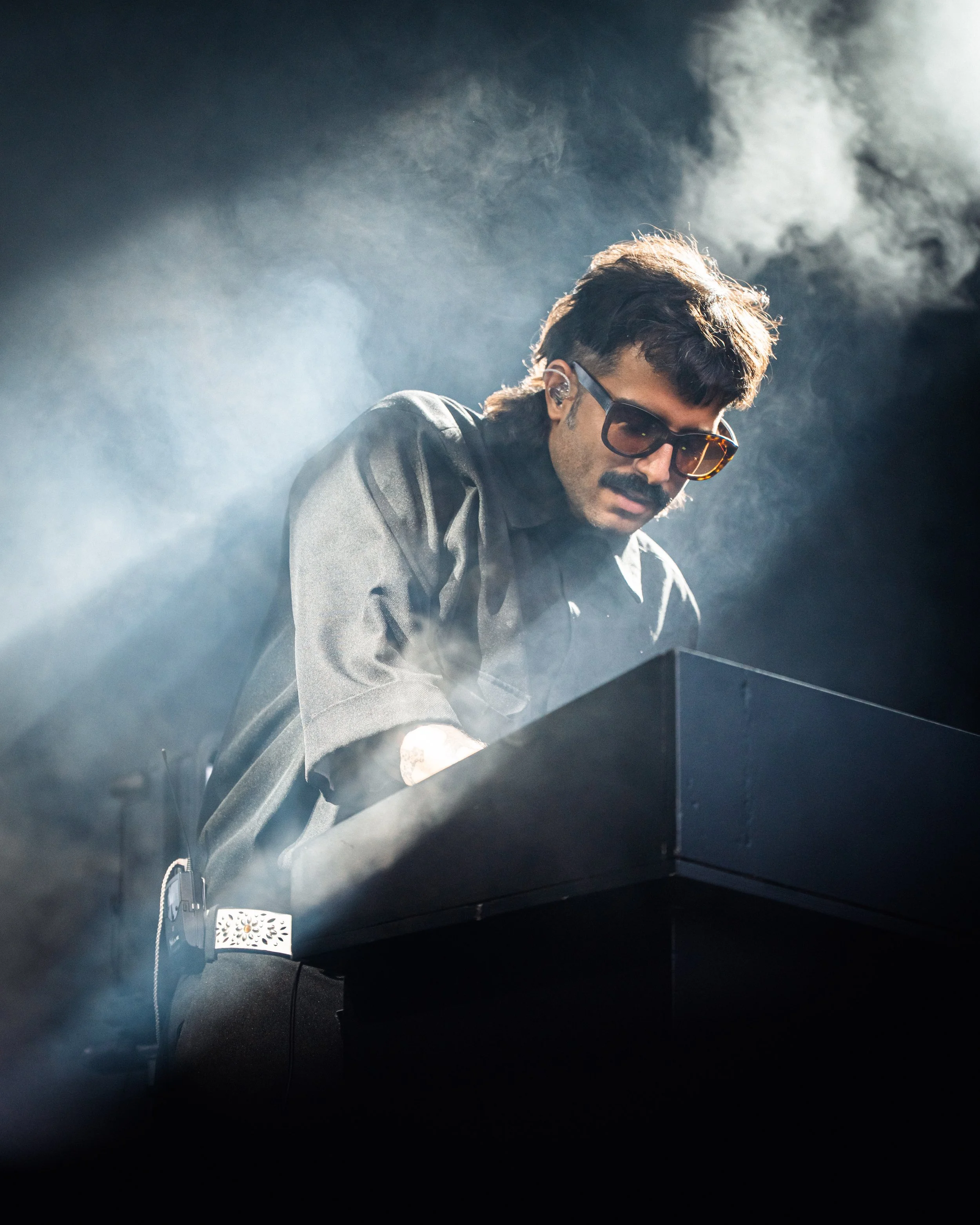 Un homme portant des lunettes de soleil joue du clavier électrique sur scène avec de la fumée autour, lumière dramatique.