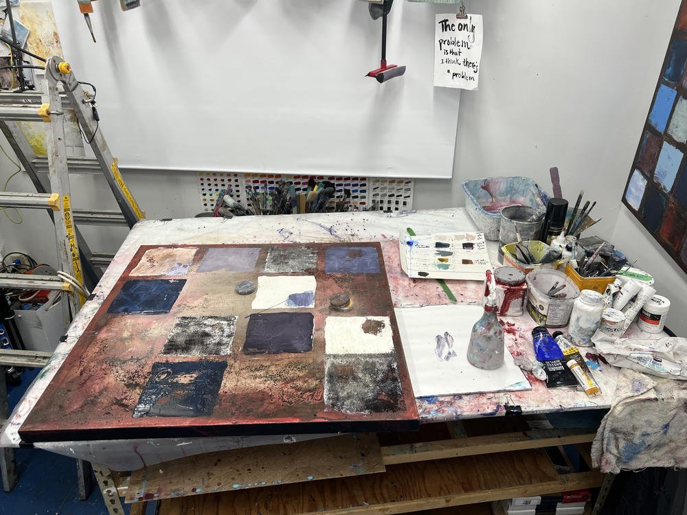 Deb Chaney's studio worktable with an in-progress painting. A handwritten not pinned to the wall above reads "The only problem is that I think there is a problem".