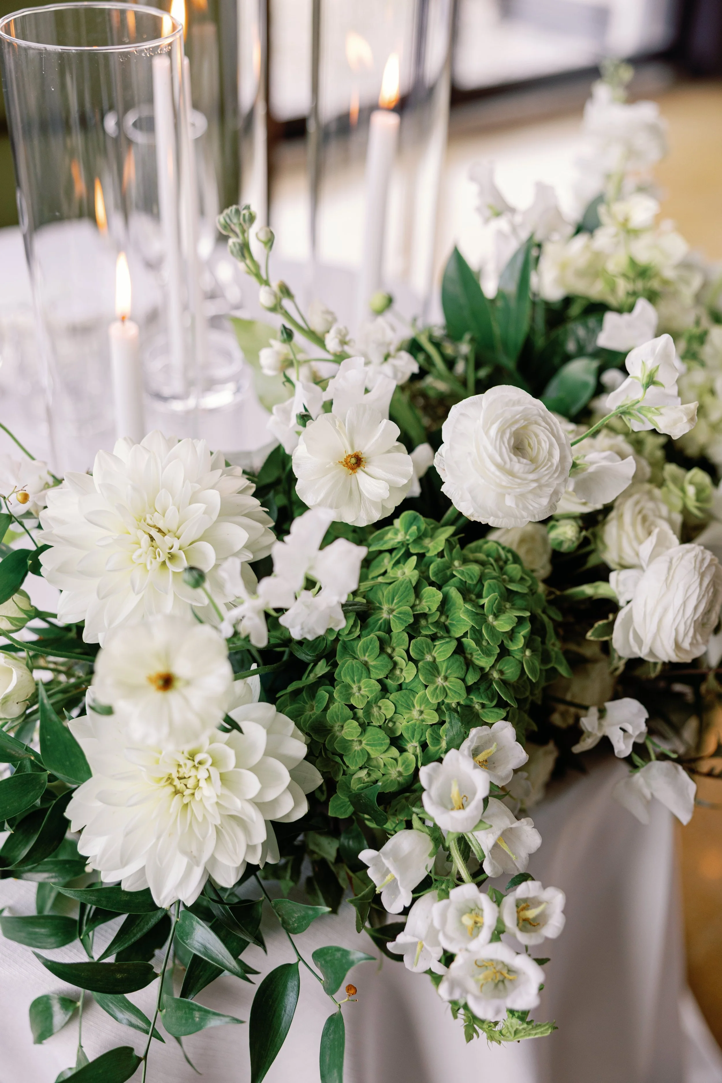 A floral centerpiece with white flowers including dahlias, ranunculus, and hydrangea, surrounded by green leaves, placed on a table with candles in the background.