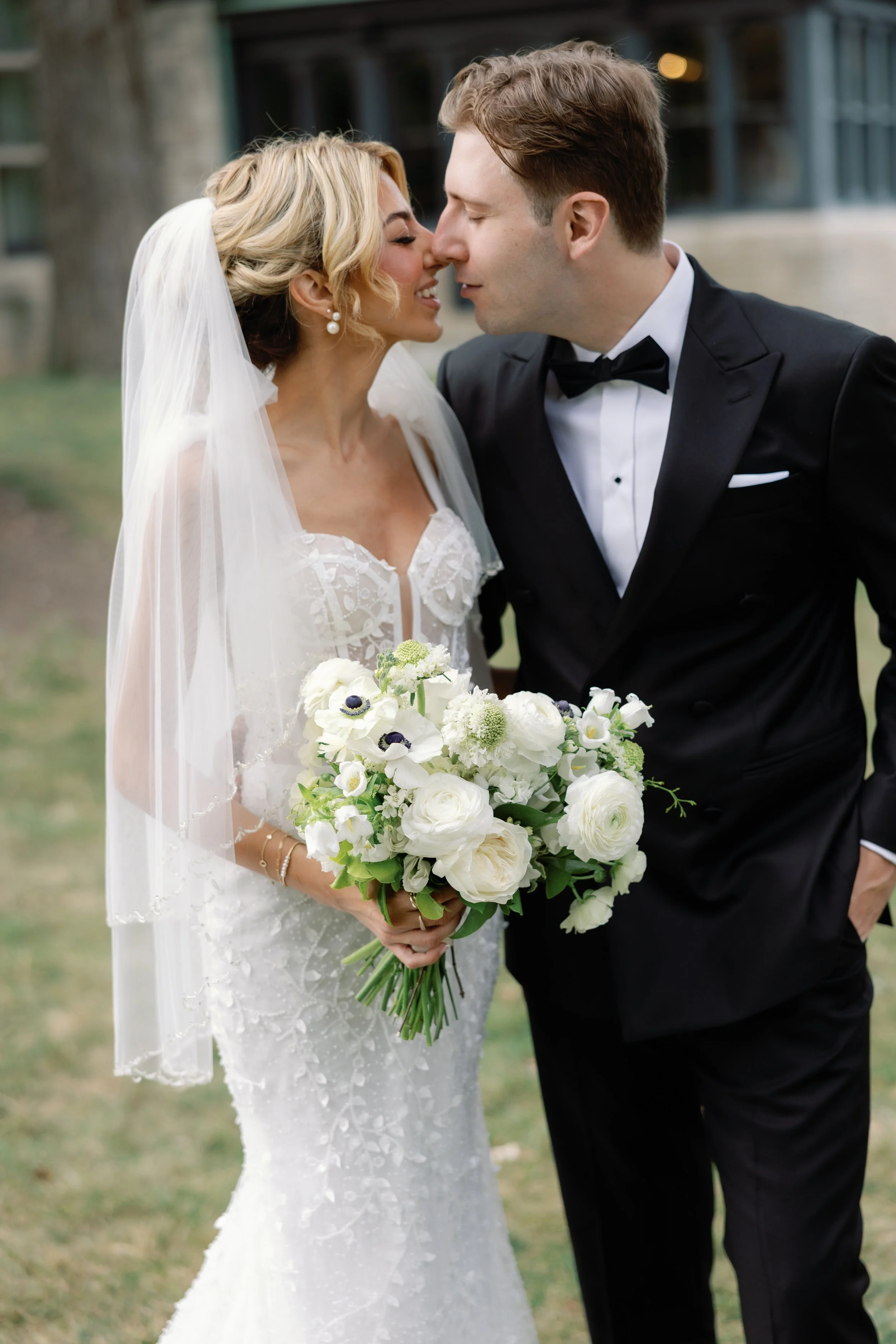 Bride and groom on their wedding day, close-up shot with wedding attire, the bride holding a bouquet of white flowers, the groom leaning towards the bride, outdoor setting.