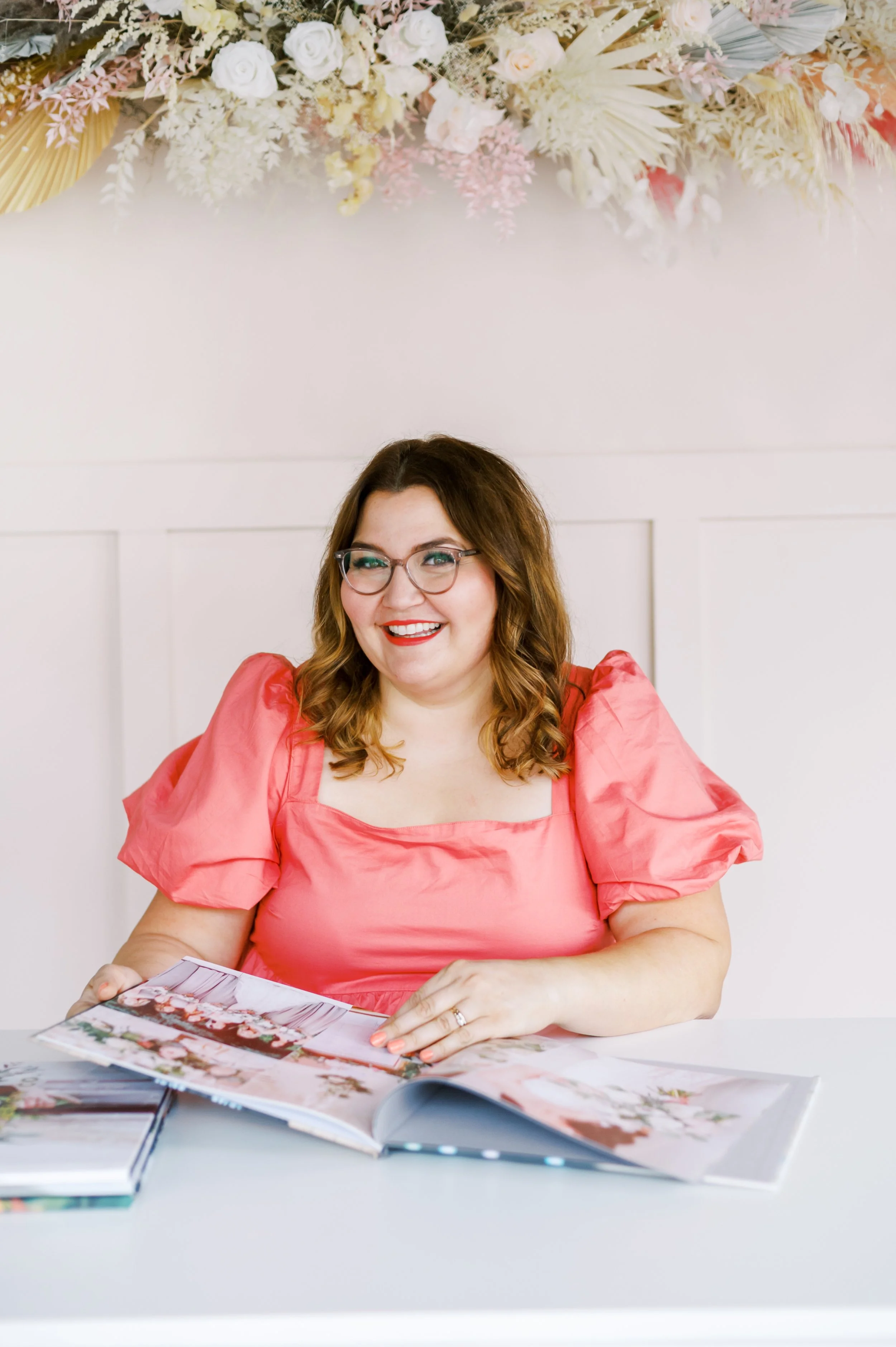 Woman with glasses and curly brown hair sitting at a white table, wearing a salmon-colored dress, smiling, looking at photo album. Floral decorations in the background.