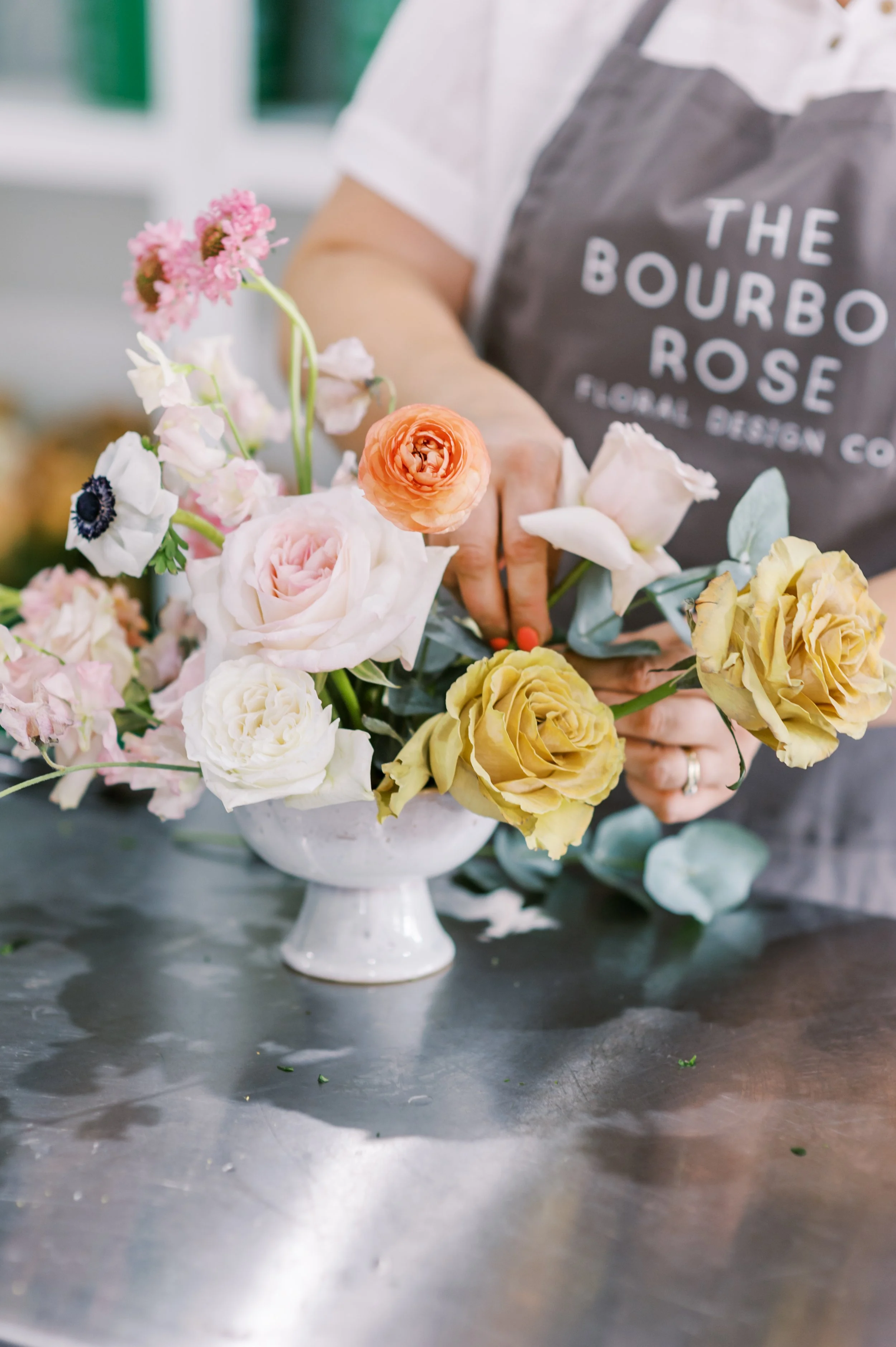 Person arranging a bouquet of mixed flowers in a white vase, wearing a apron that reads 'The Bourbon Rose Floral Design Co.'