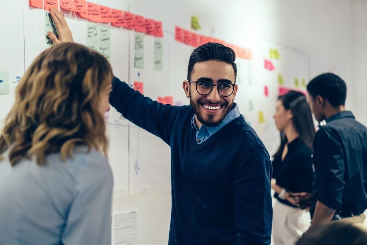 Learning strategies: A man happily points to a Post-It note on a wall full of them. They each have written notes and are organized in lines.