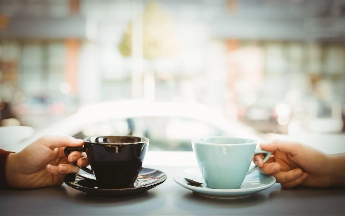 Two mugs sit on a table with two people holding the handles.