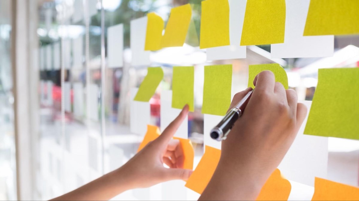Blank Post-It notes on a window. One person poises their hand to write and another points.