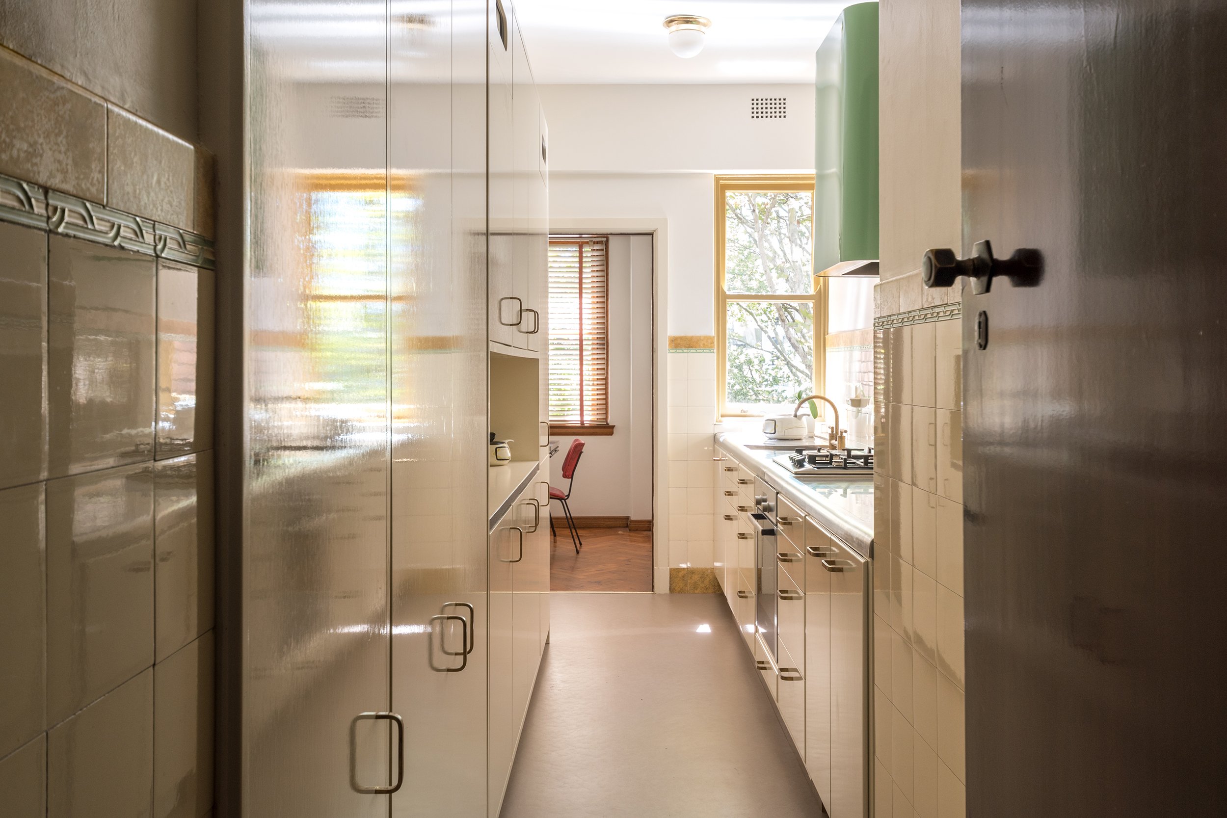 A view down the revamped galley kitchen to the dining room, highlighting the original 1938 wall tiles paired with hand-painted joinery in colours chosen to complement them. The all-new cabinetry conceals appliances, while an Art Deco-inspired shroud