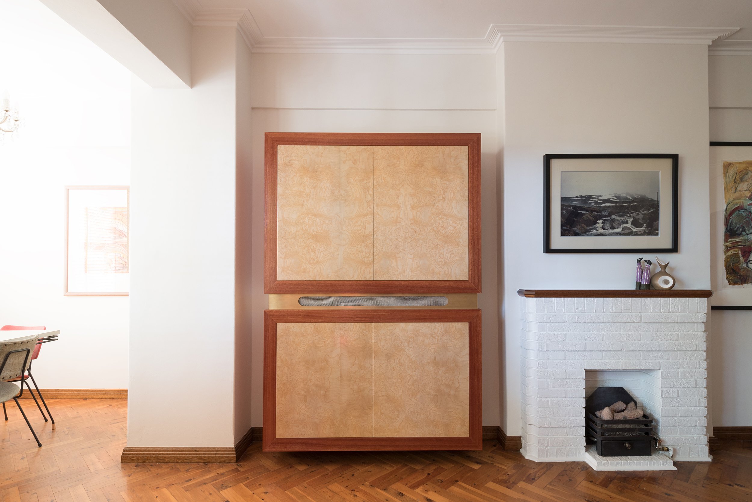 A custom media cabinet designed to conceal the TV and fridge poking through from behind, crafted with a mix of timber veneers, including burl, that complement the original timbers in this Art Deco apartment. The cabinet functions as a TV and stereo