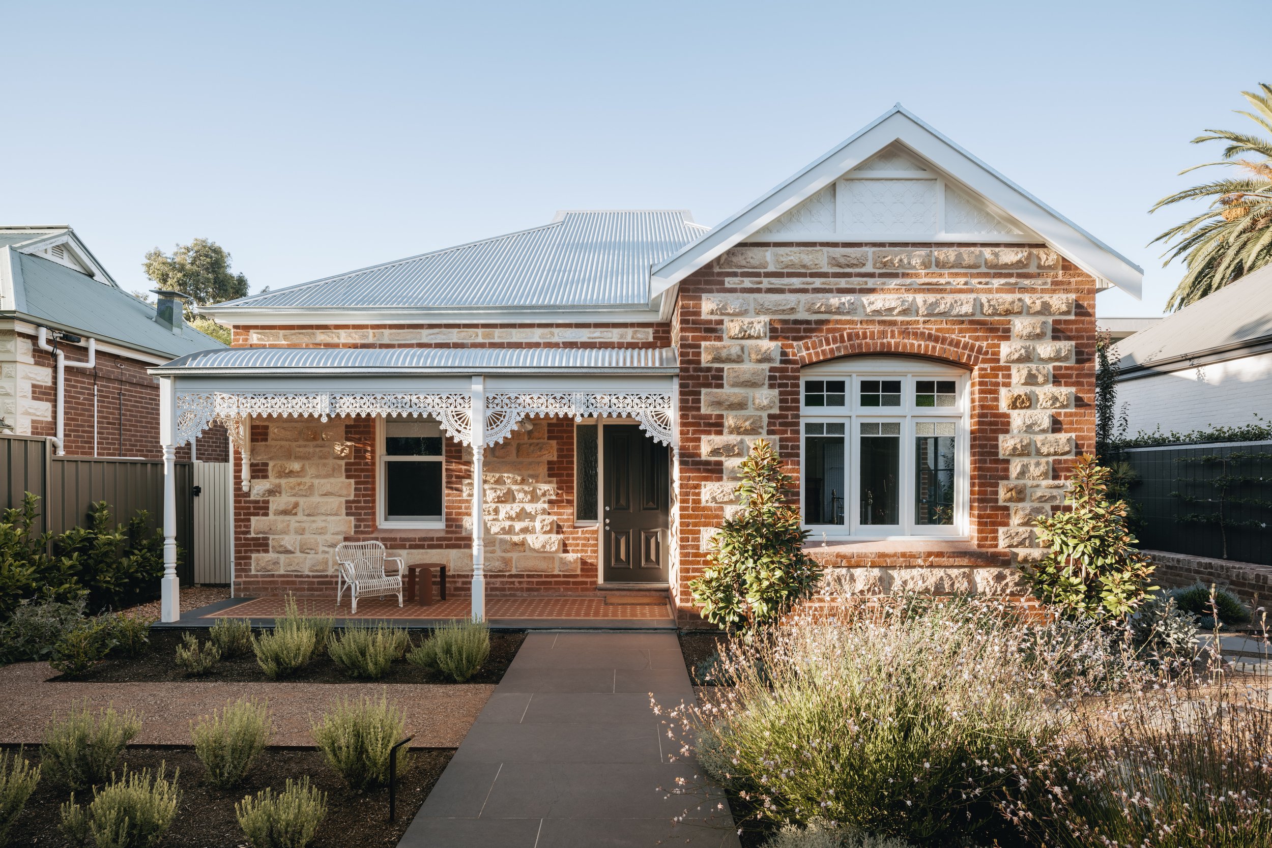 Front view of a restored Adelaide villa with Adelaide Hills sandstone, featuring brick coining, repointed masonry, and a rebuilt verandah with original restored wrought iron lacework and timber windows. The contemporary cottage garden complements th