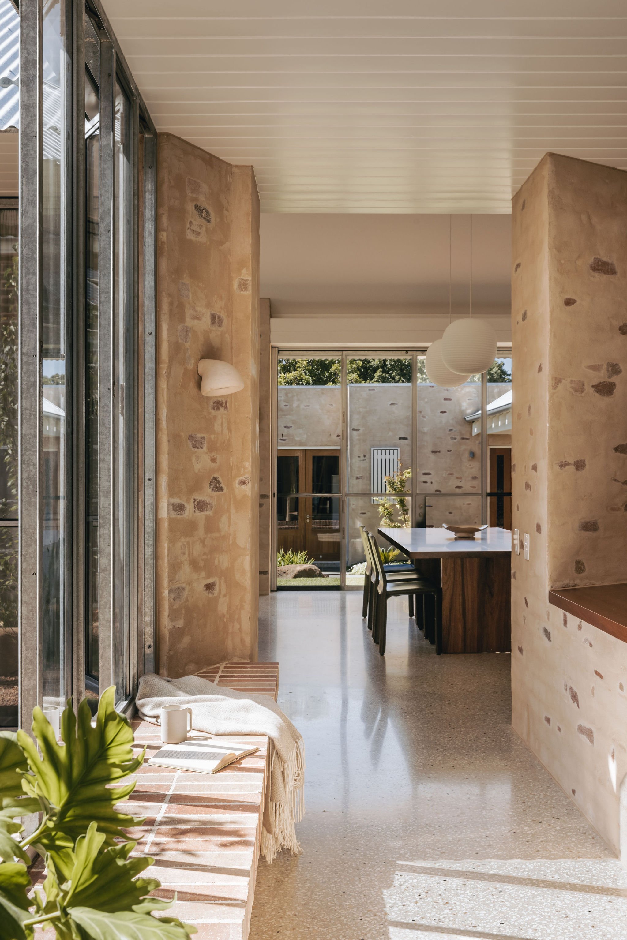 View down the hallway into the new living space, showcasing large brick window sill, galvanised windows, and a dining area featuring a locally made timber table, glass pendant lanterns, and bagged brickwork. Polished concrete floors and matchboard t