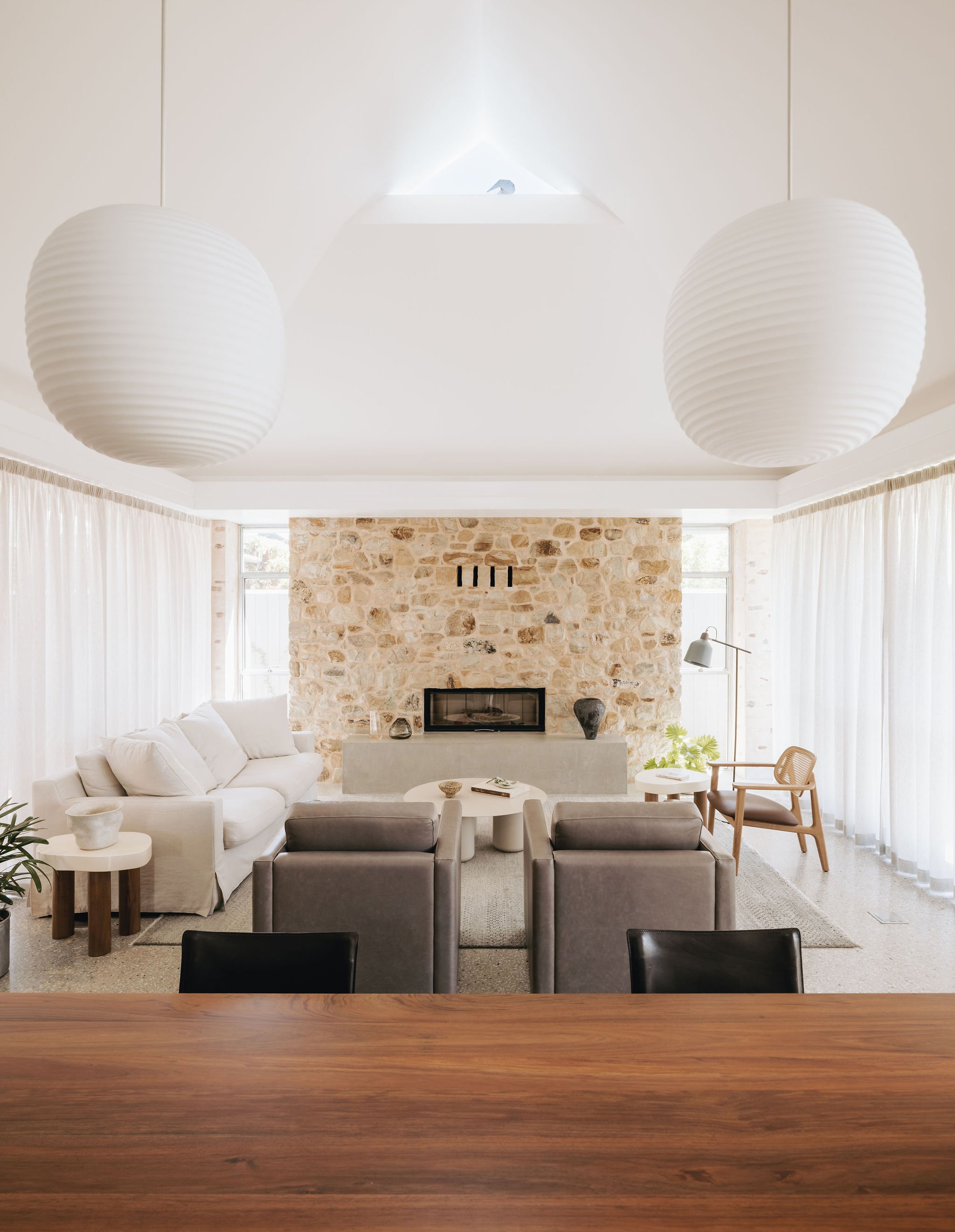 Angle across the dining table to the living room, featuring a timber dining table with glass lanterns and vintage dining chairs. A solid concrete hearth and sandstone chimney breast anchor the space, complemented by custom locally made leather tub c