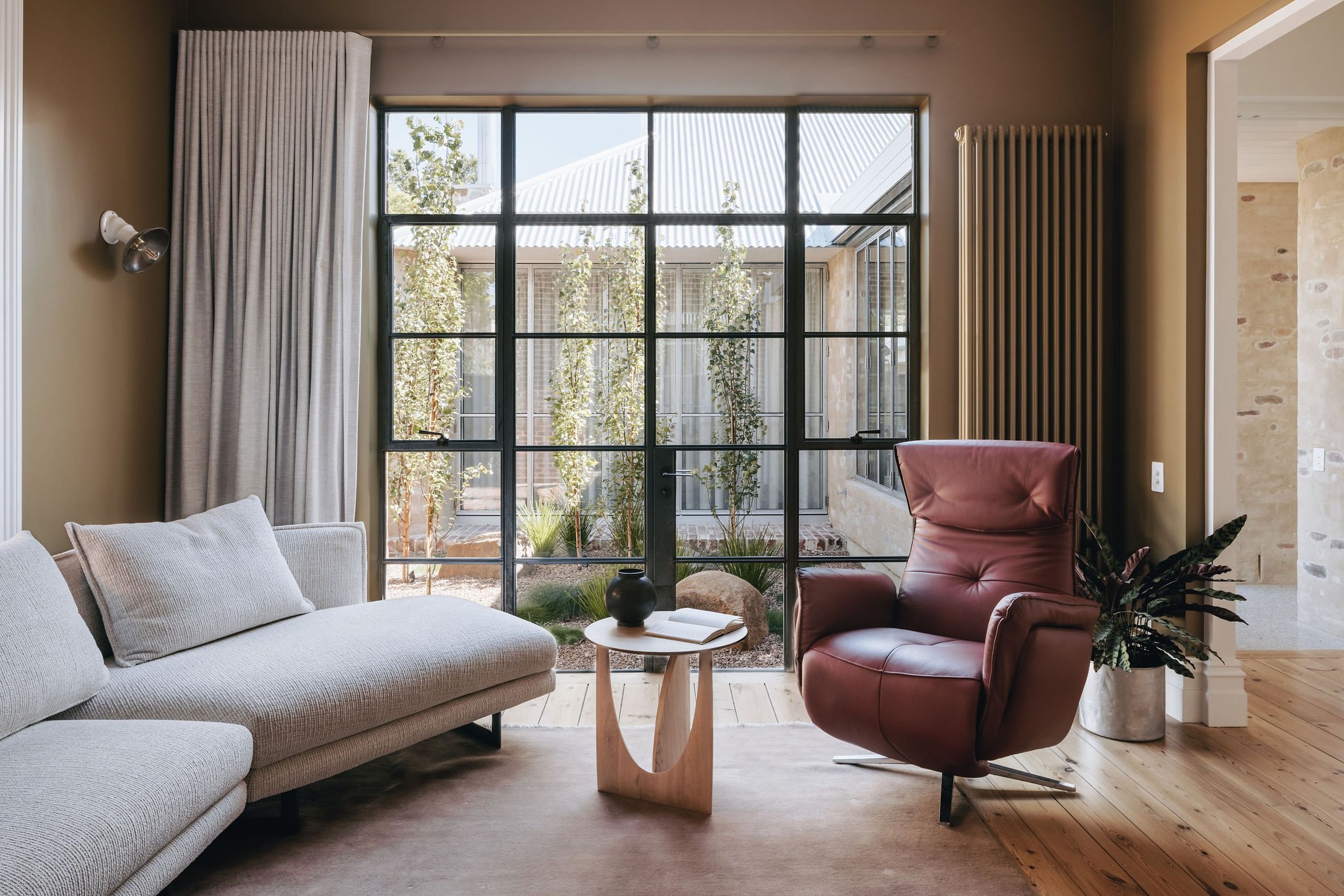 View from the original part of the house into the newly created courtyard, showcasing original timber floorboards, a custom-coloured radiator, and galvanised windows. The deep, warm Dulux Research wall colour complements the space, while the courtya