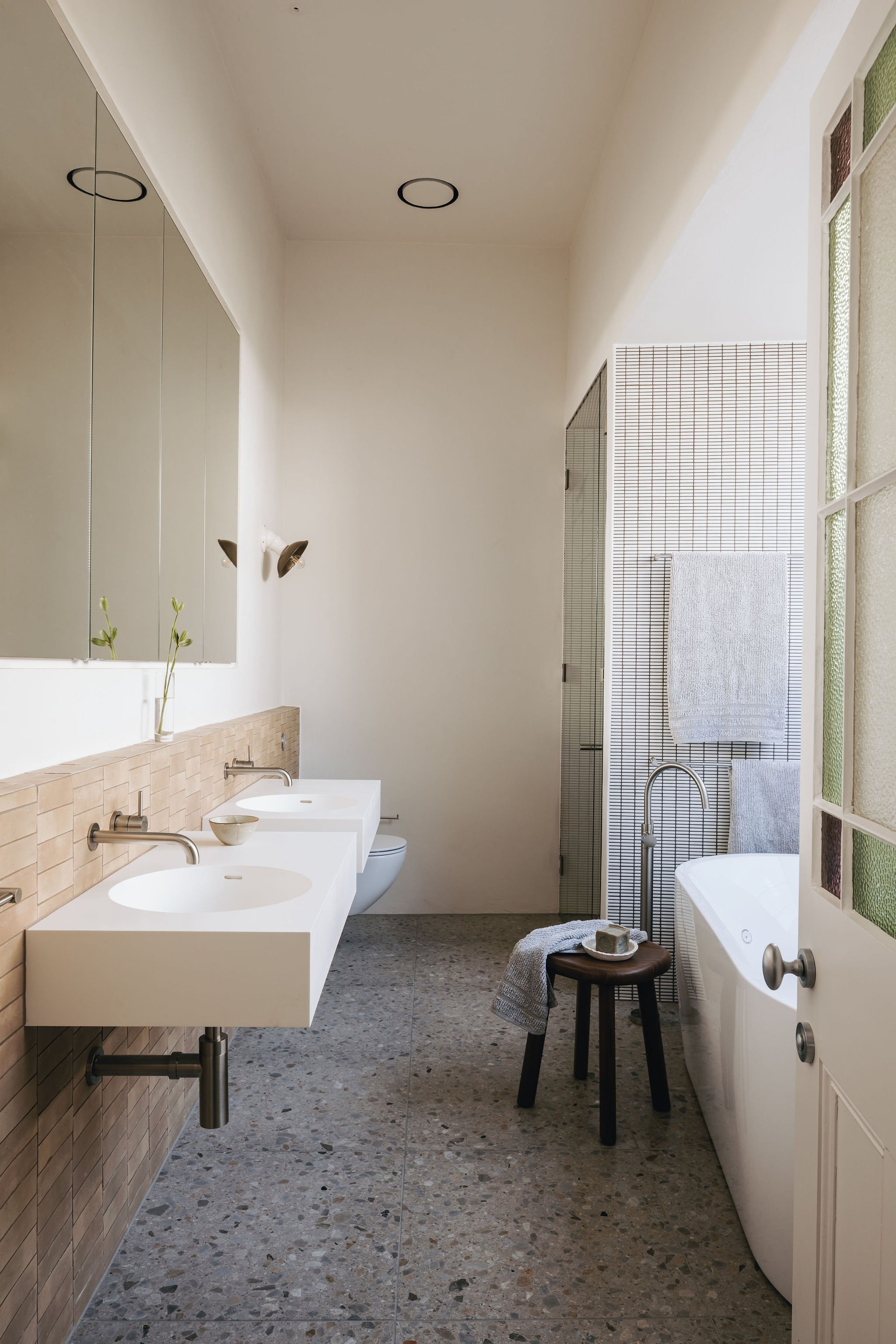 A glimpse of the ensuite space, featuring earthy terracotta wall tiles that complement the terrazzo tile flooring. Recessed mirror cabinets, wall-hung sanitaryware, and Venetian plaster walls create a clean, modern aesthetic. The steam shower enhanc
