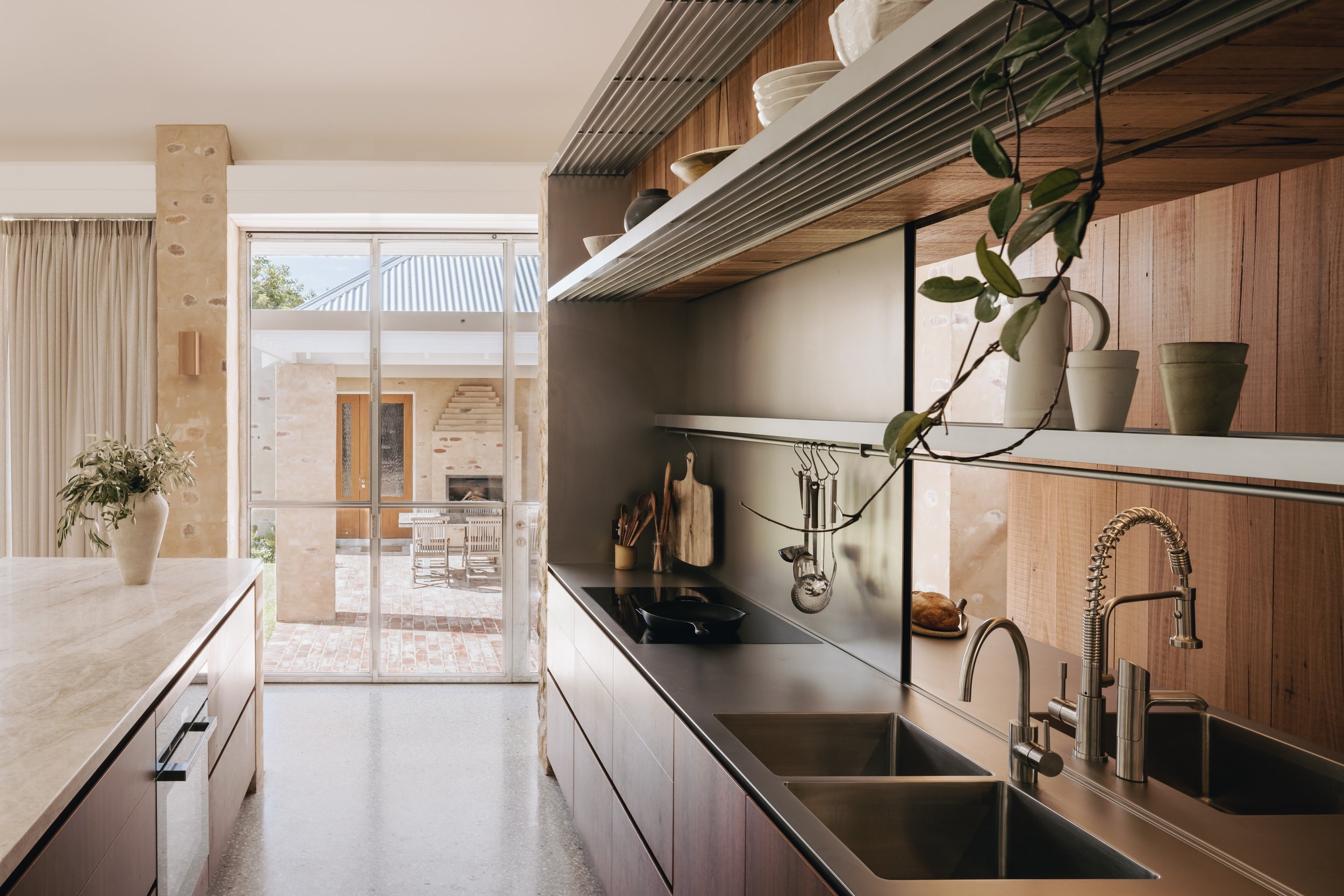 A detailed view of the kitchen highlights the stainless steel work, including a bench with integrated sinks and an operable sliding stainless steel splashback that opens to reveal the pantry. Shelves made from stainless steel bars add a functional i